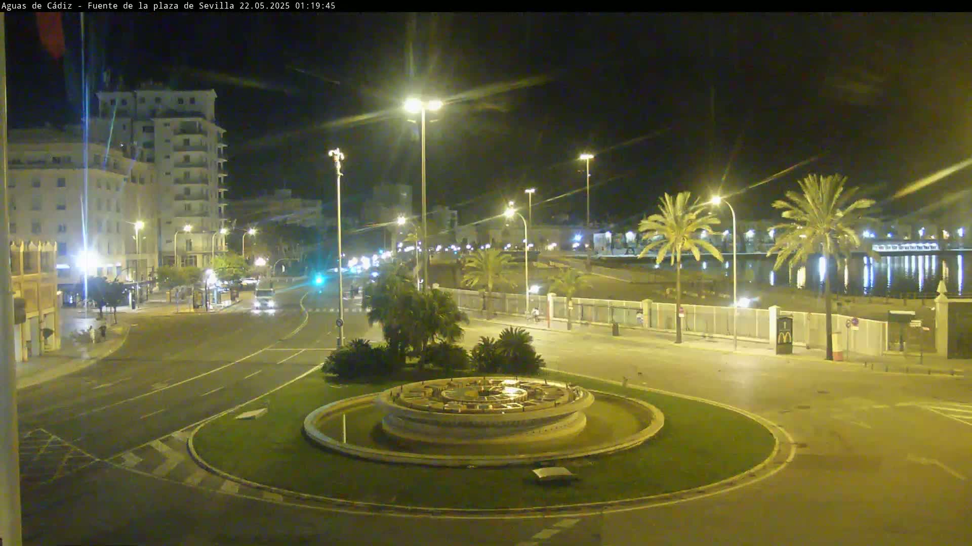 A nighttime view of a circular plaza with a fountain in the center, surrounded by palm trees, buildings, and roadways.
