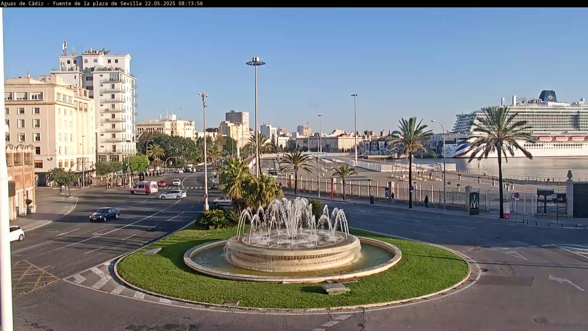 A city roundabout with a central fountain is shown, with a cruise ship visible in the harbor in the background under a clear, sunny sky.