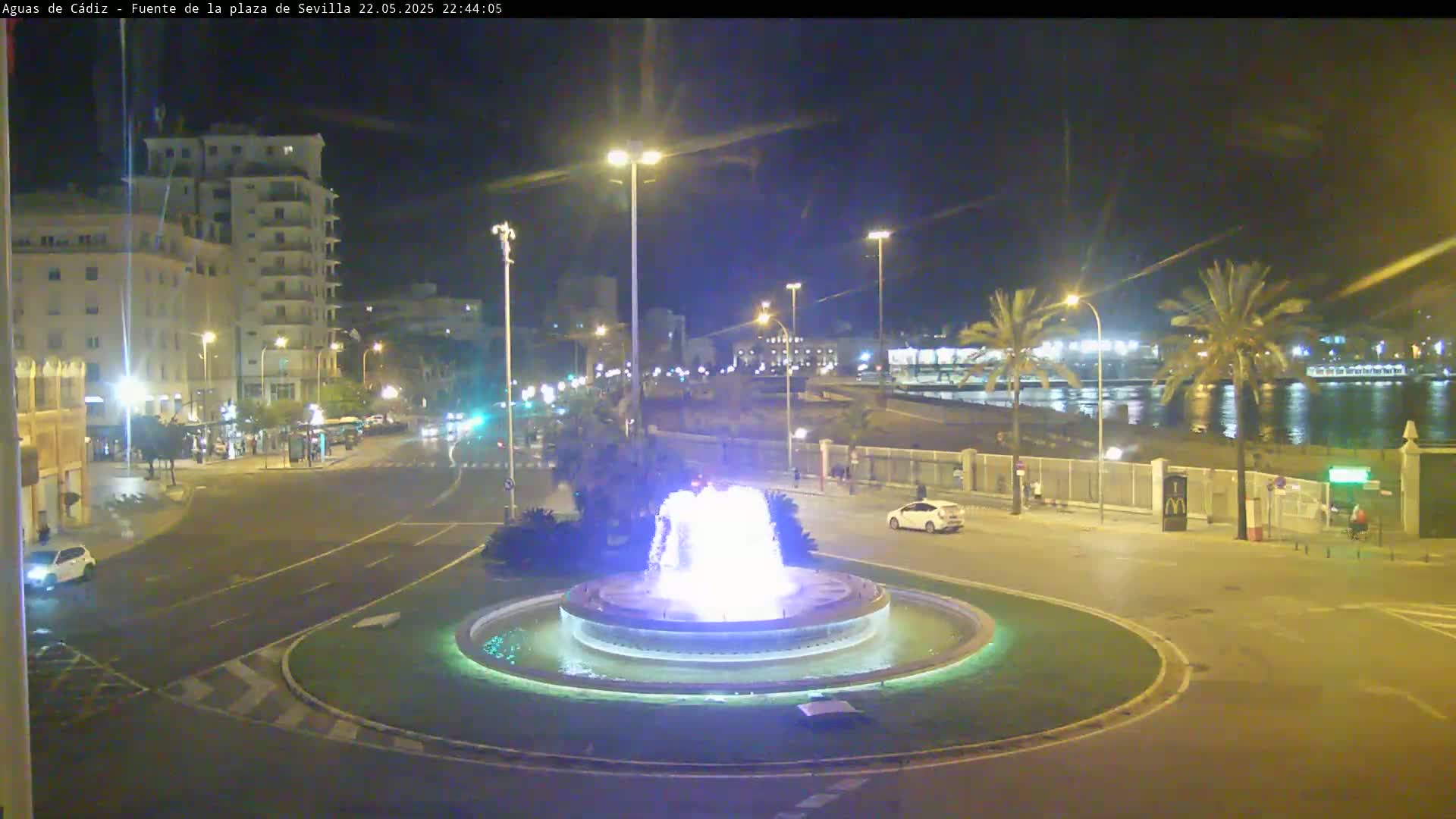 A nighttime view of a city square with a brightly lit fountain at its center, bordered by buildings, palm trees, and a waterfront.
