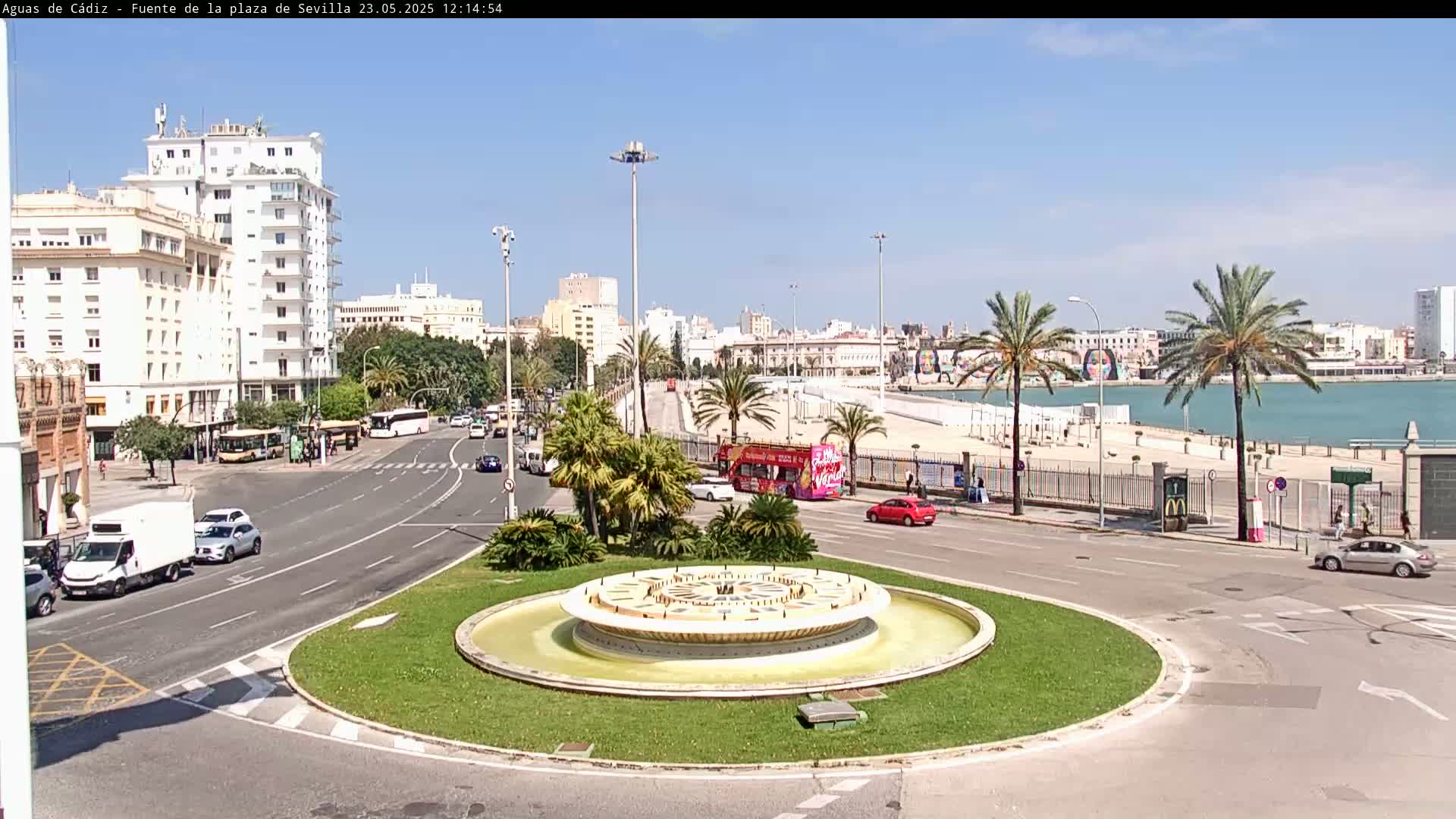 A circular fountain sits in a traffic circle in a city by a body of water on a sunny day.