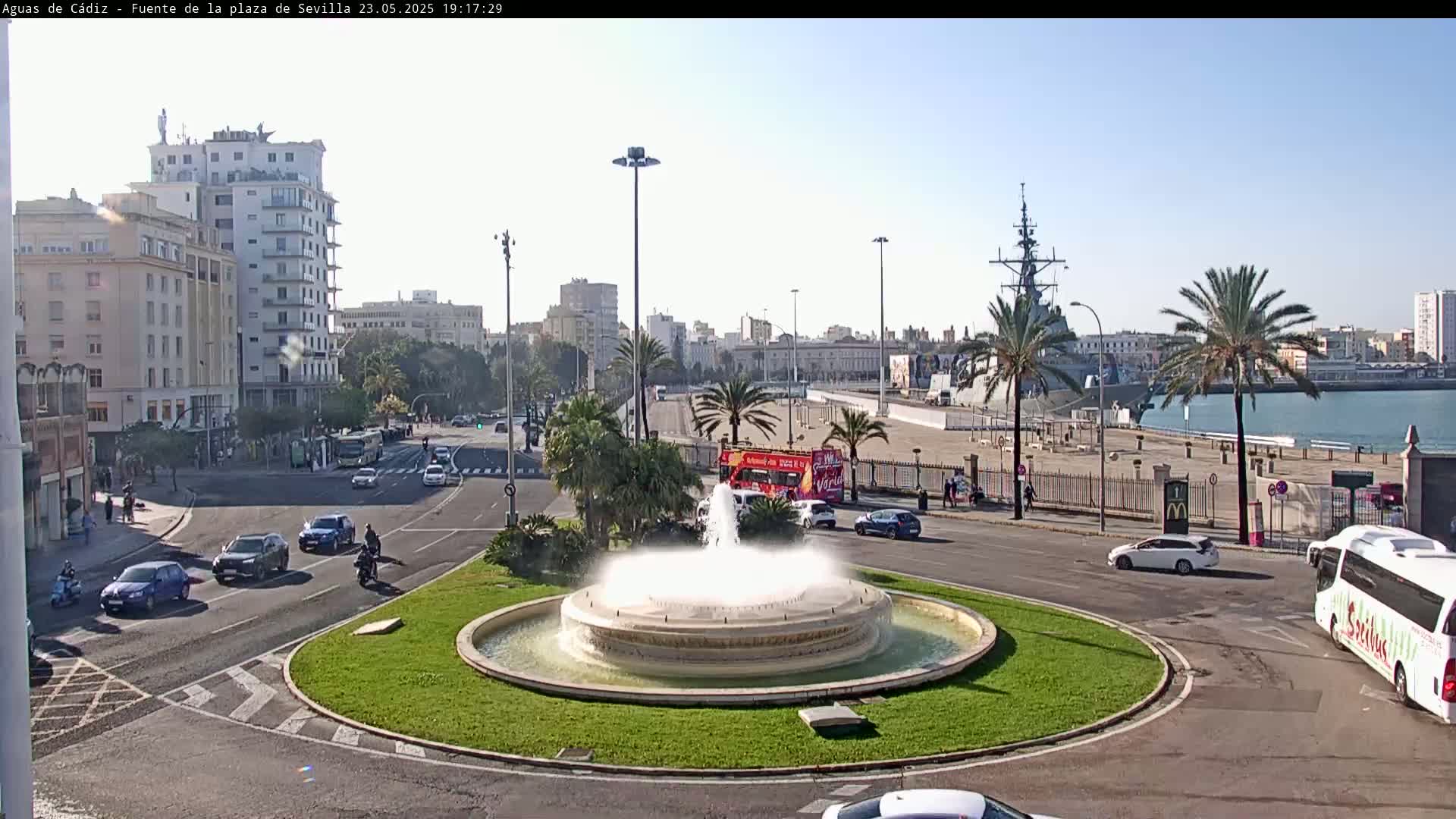 A city roundabout with a fountain in the center, surrounded by cars, palm trees, and buildings, overlooks a harbor with a docked warship under a mostly clear sky.