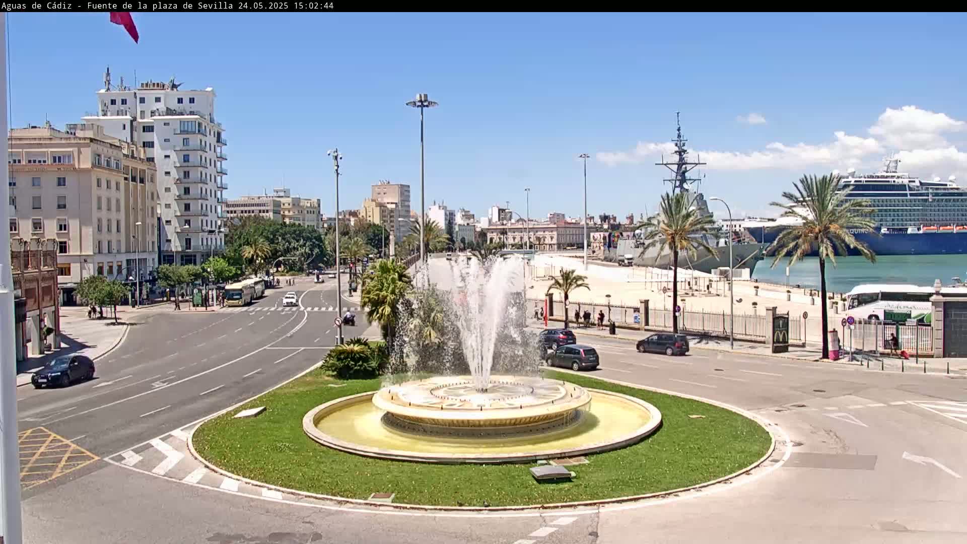 A large fountain in a traffic circle is the focal point of a sunny city scene overlooking a harbor with a docked cruise ship and warship.
