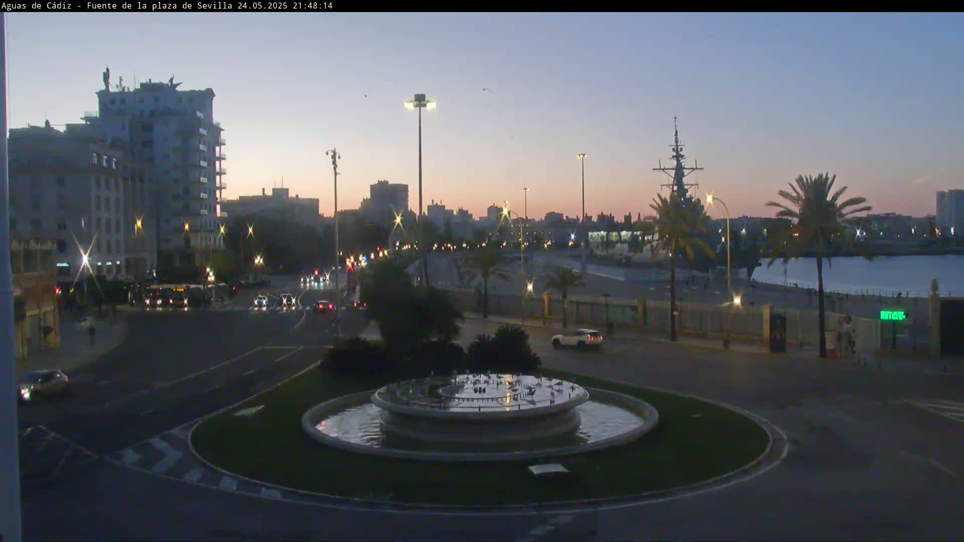 A city roundabout with a fountain at its center is surrounded by buildings, palm trees, and a waterfront with a large ship visible at dusk under a clear sky.