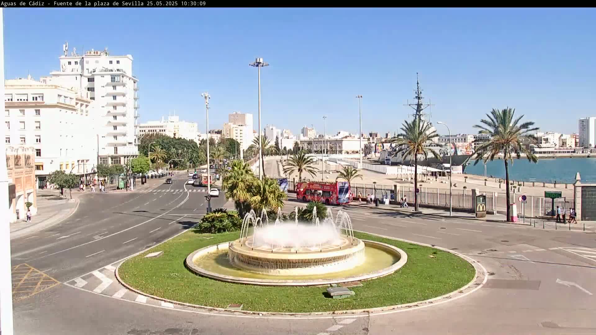 A large fountain in a city roundabout, with palm trees, buildings, and a body of water with a ship in the background, under sunny skies.