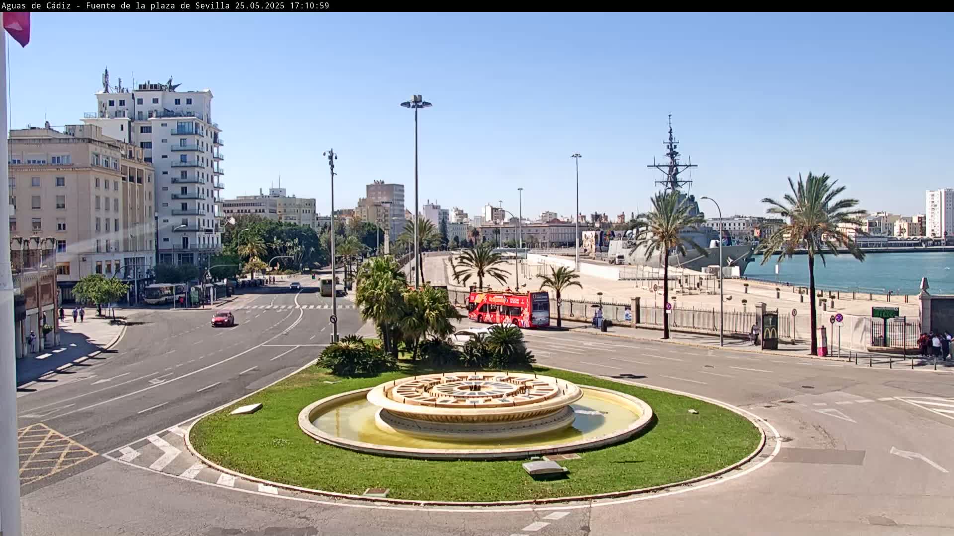 A circular fountain sits in the center of a traffic circle, with a city skyline, palm trees, a red double-decker bus, and a docked warship visible in the background under a clear, sunny sky.