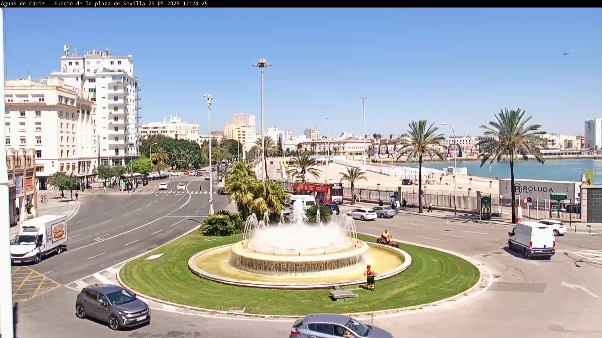 A large fountain in a traffic circle is surrounded by palm trees, buildings, and cars under a clear blue sky.