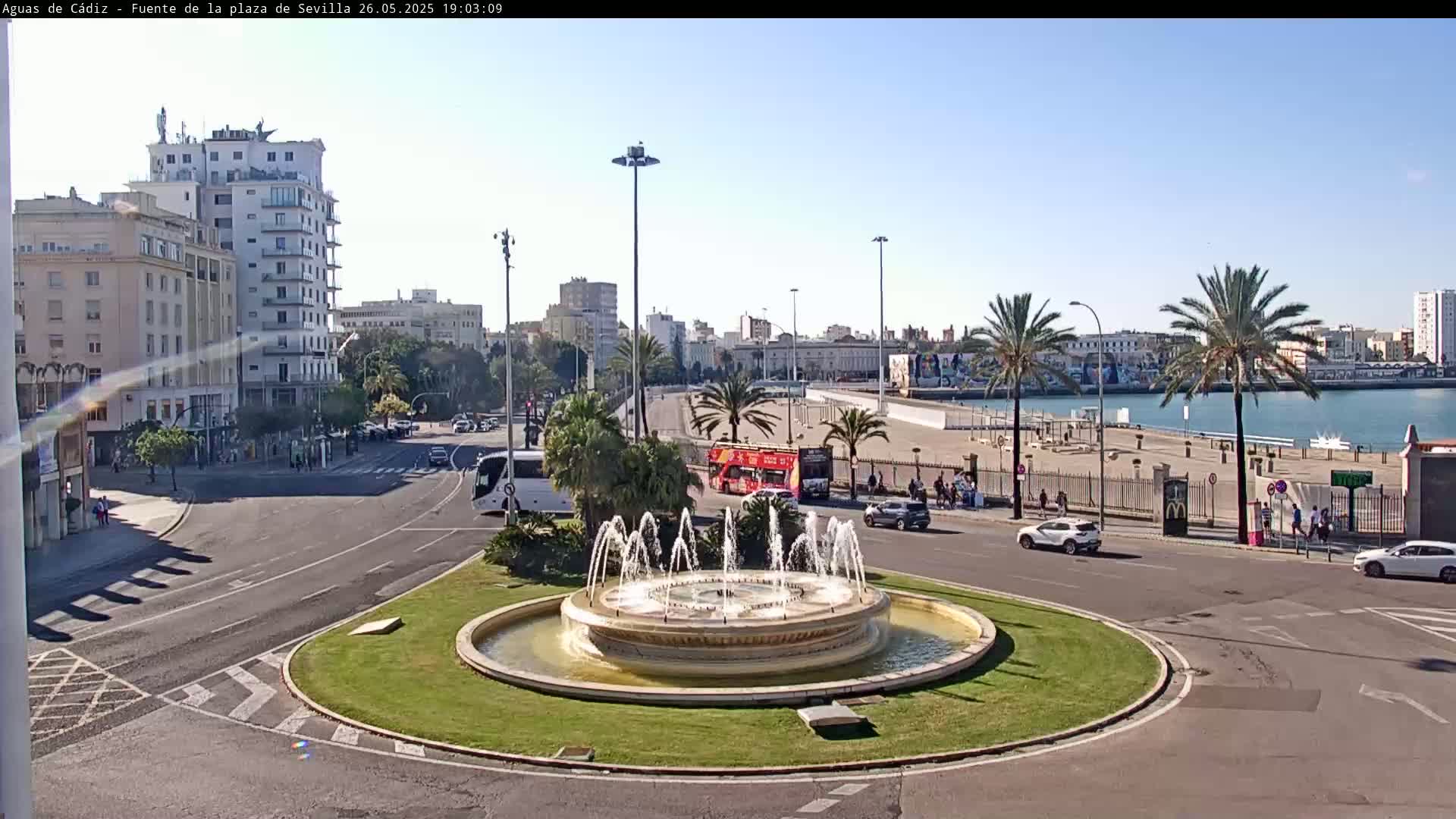 A circular fountain is the center of a city traffic circle near a waterfront, under a sunny sky.