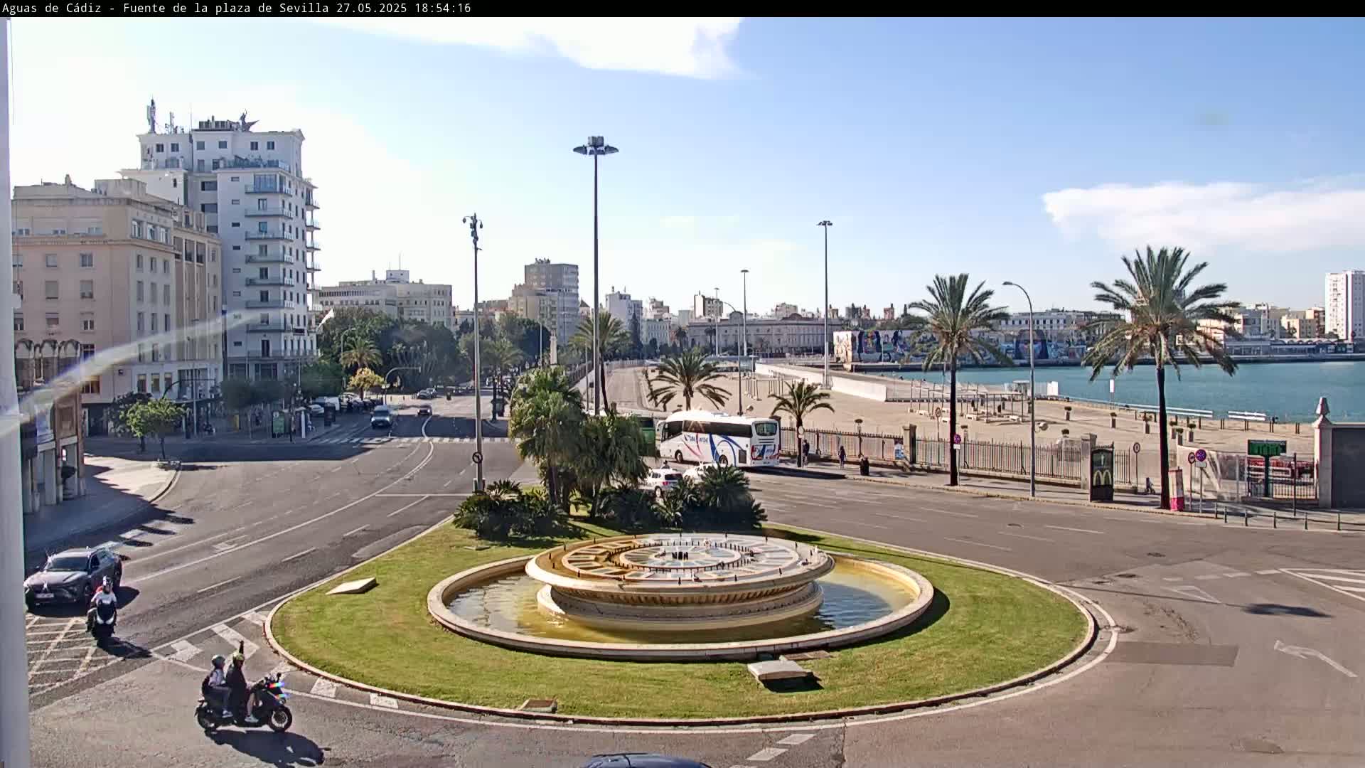 A city roundabout with a fountain in the center, bordered by palm trees and buildings, overlooks a waterfront under a partly cloudy sky.