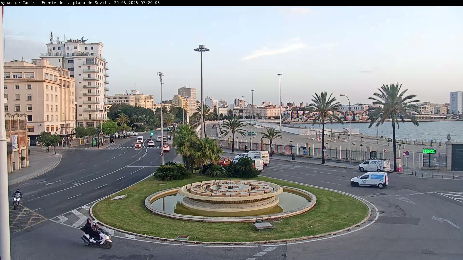 A roundabout with a central fountain is situated near a waterfront, with several buildings and palm trees lining a roadway under a clear, bright sky.