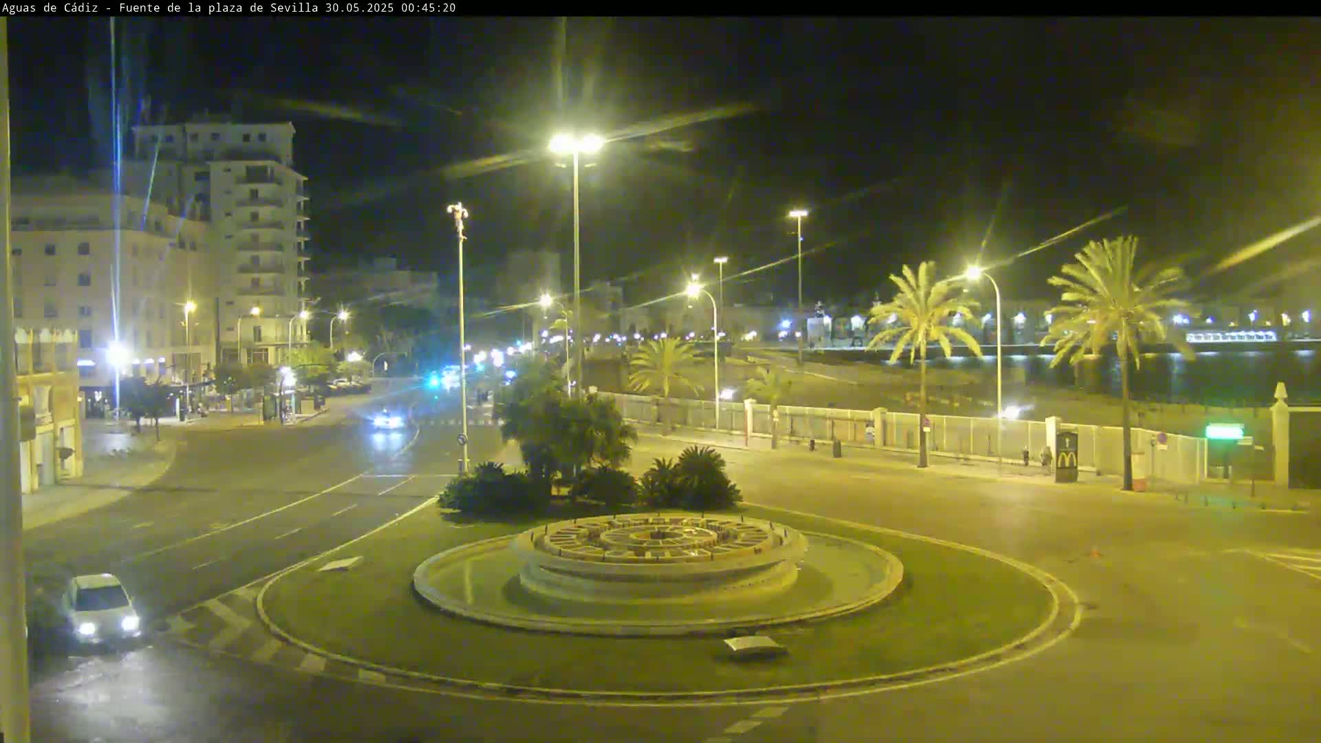 A nighttime view of a roundabout with a fountain at its center, surrounded by palm trees, buildings, and roads with sparse traffic.