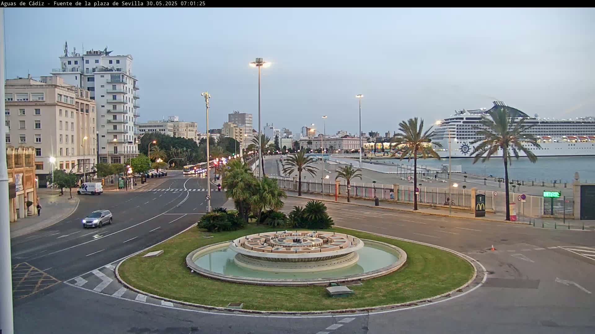 A city waterfront scene at dusk shows a circular fountain in the foreground, a roadway with sparse traffic, buildings lining the street, palm trees, and a large cruise ship docked in the harbor.