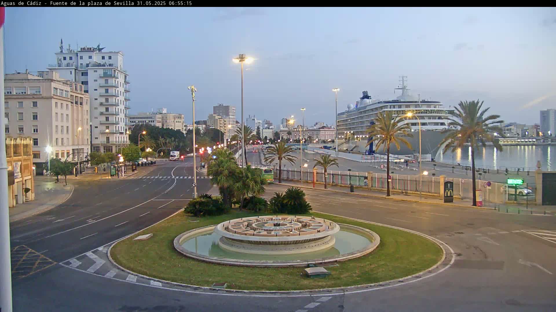 A large cruise ship is docked at a harbor near a city with a circular fountain in the foreground, under a twilight sky.