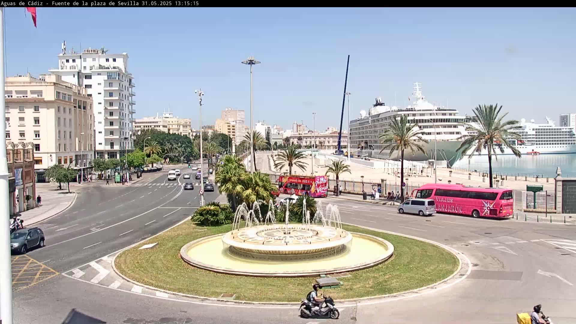 A city roundabout with a large fountain in the center features several vehicles, including buses and cars, near a harbor with docked cruise ships on a sunny day.