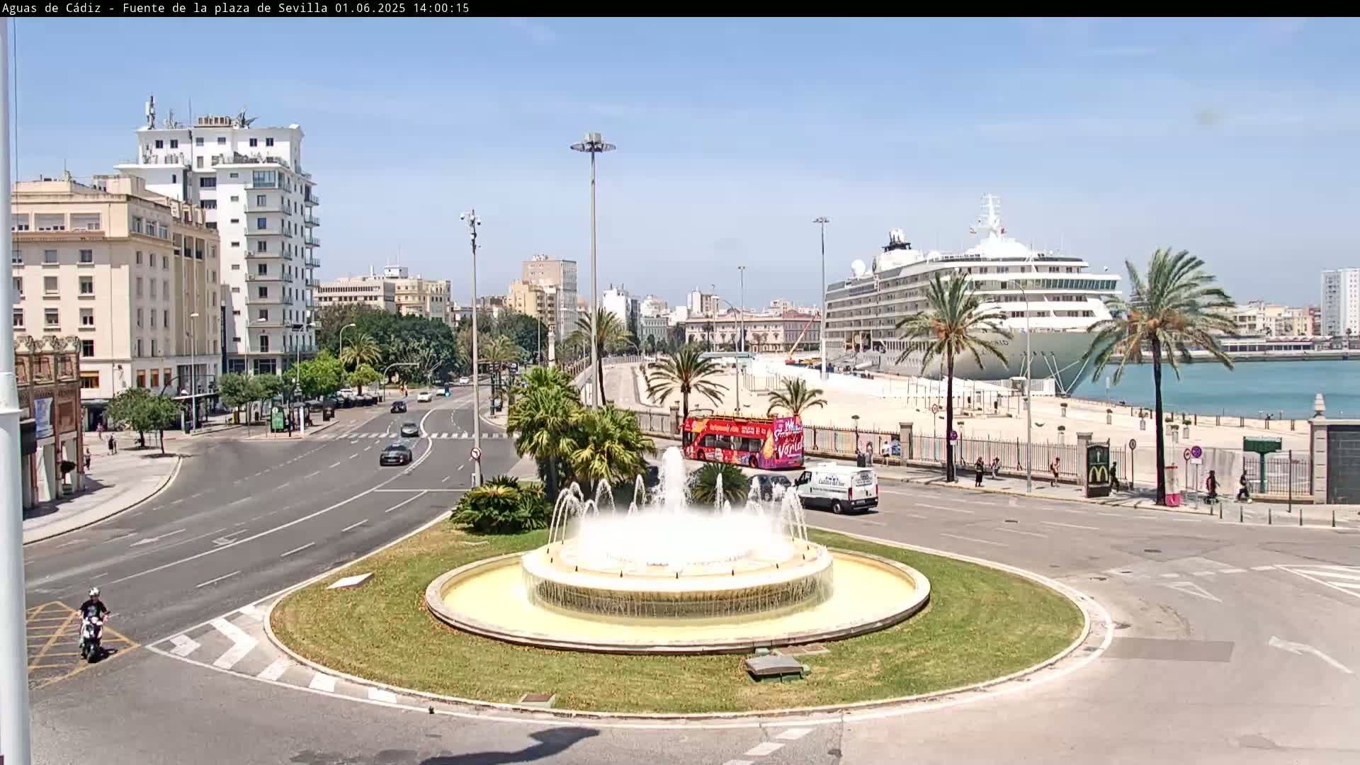 A large cruise ship is docked at a harbor near a city with a fountain in a traffic circle, under a clear sunny sky.