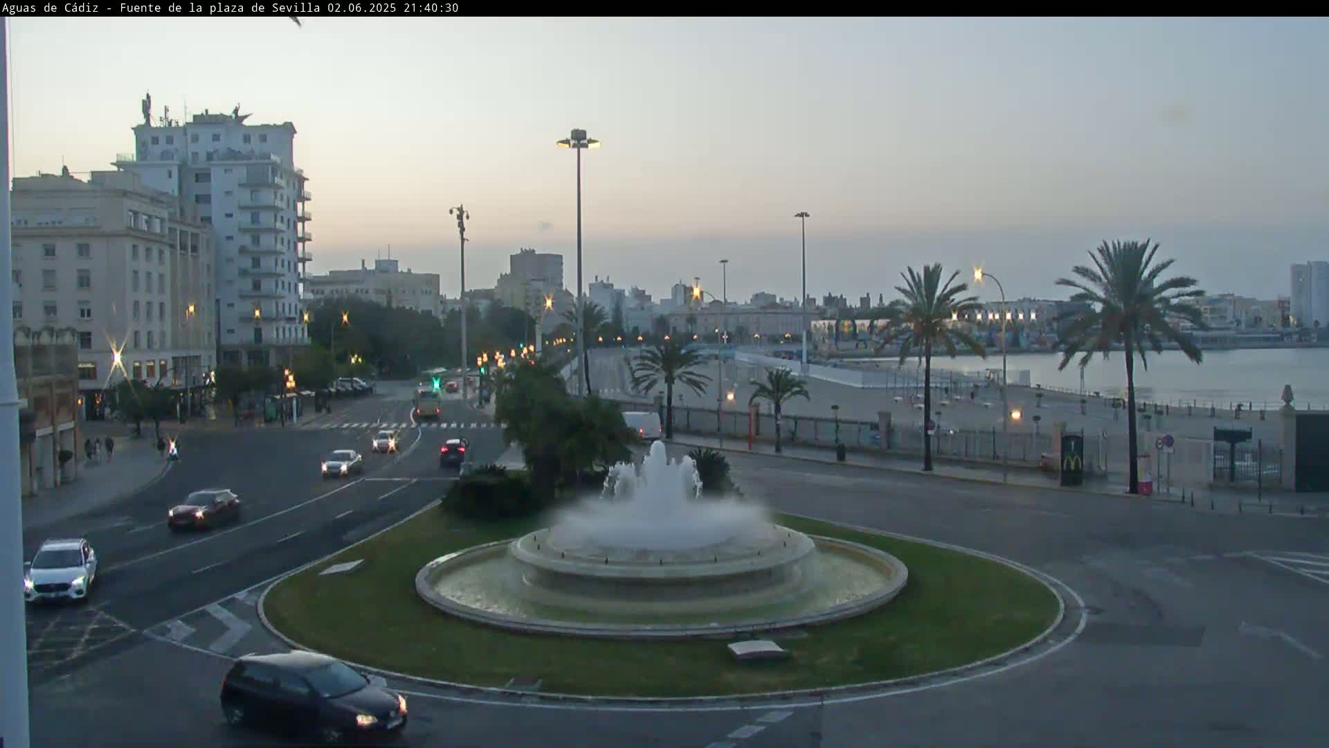 A city street scene at twilight shows a fountain in a roundabout, with cars driving on the streets and buildings and palm trees visible in the background.
