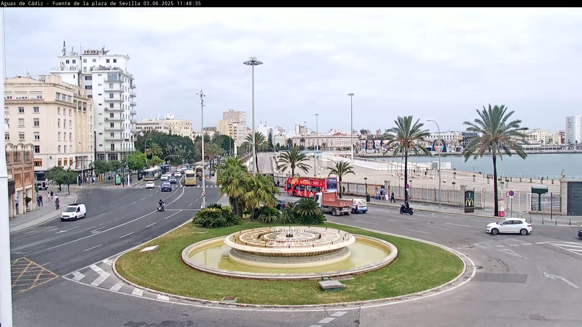 A circular fountain sits in the center of a traffic circle, surrounded by city buildings, palm trees, and vehicles under a cloudy sky.