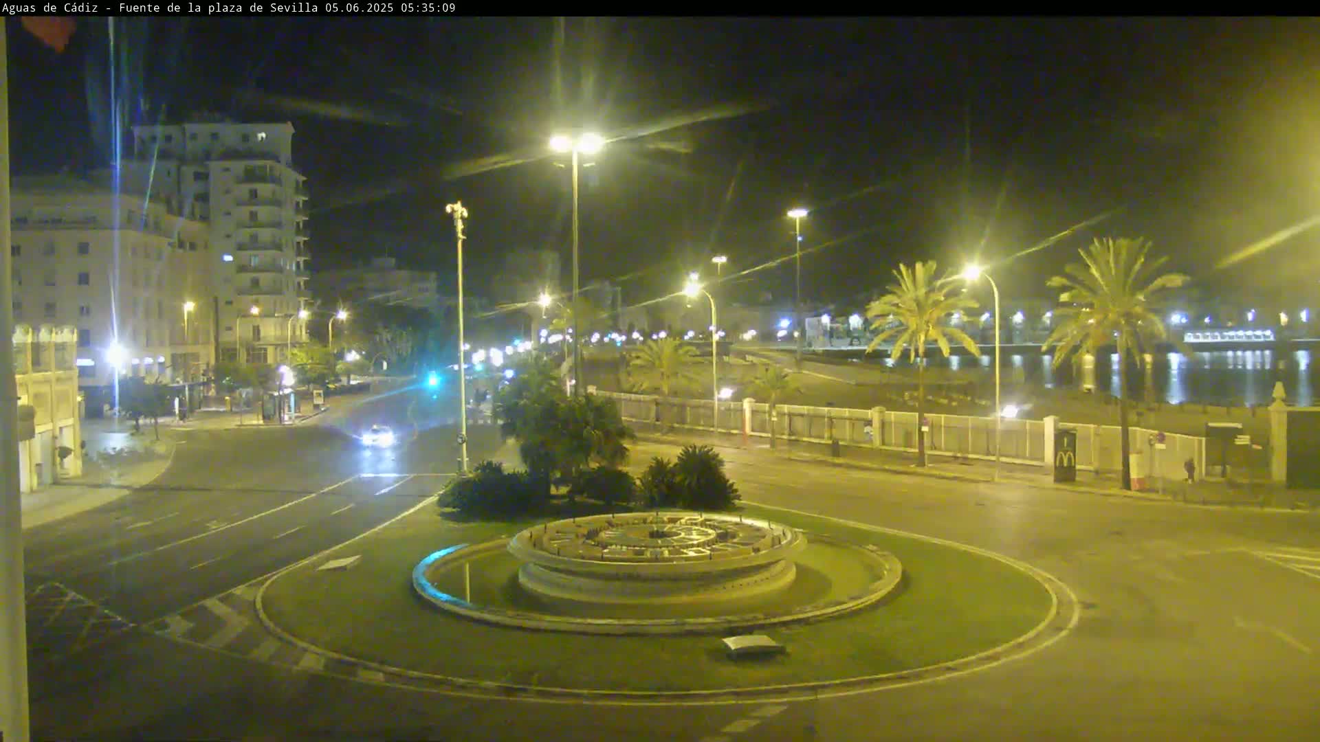 A nighttime view of a city roundabout with a fountain in the center, bordered by palm trees, buildings, and a waterfront.
