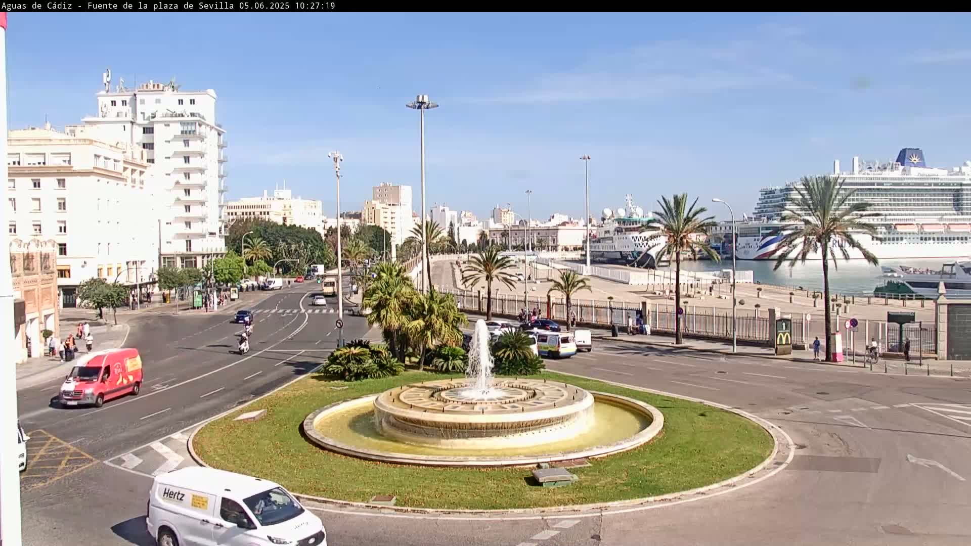 A city roundabout with a central fountain, surrounded by palm trees and buildings, overlooks a harbor with a large cruise ship on a sunny day.