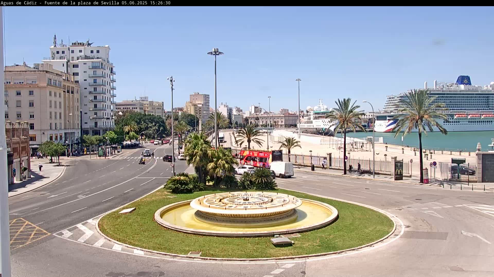 A circular fountain sits in the center of a traffic circle, with city buildings, palm trees, and a large cruise ship visible in the background on a sunny day.