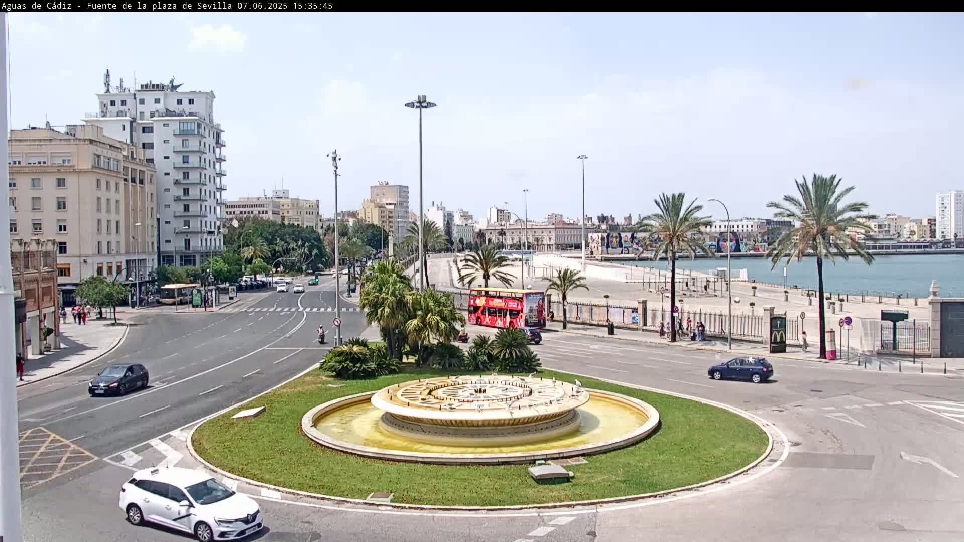 A circular fountain sits in the center of a traffic circle, surrounded by palm trees, buildings, and a waterfront with a red double-decker bus visible in the distance, under a sunny sky.