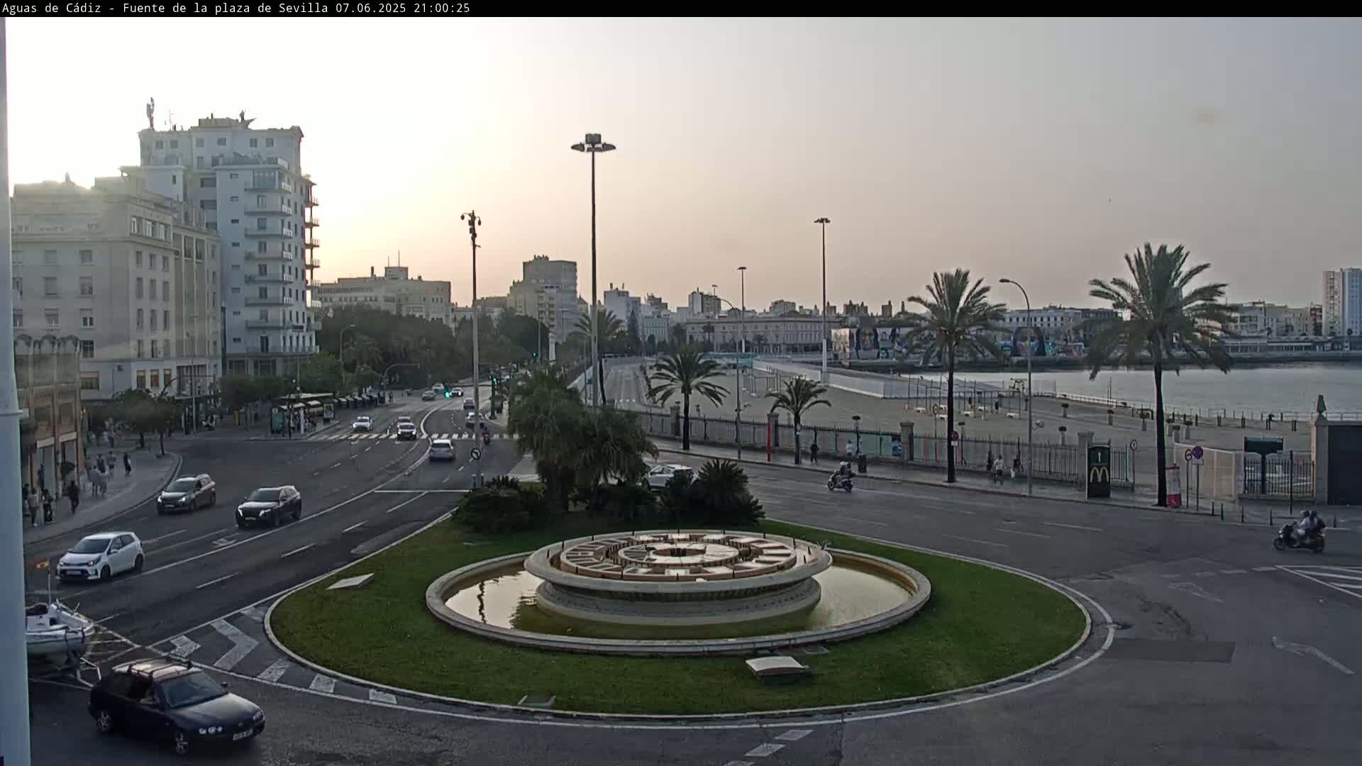 A city roundabout with a fountain in the center, surrounded by roads with cars, buildings, palm trees, and a body of water in the background under a twilight sky.