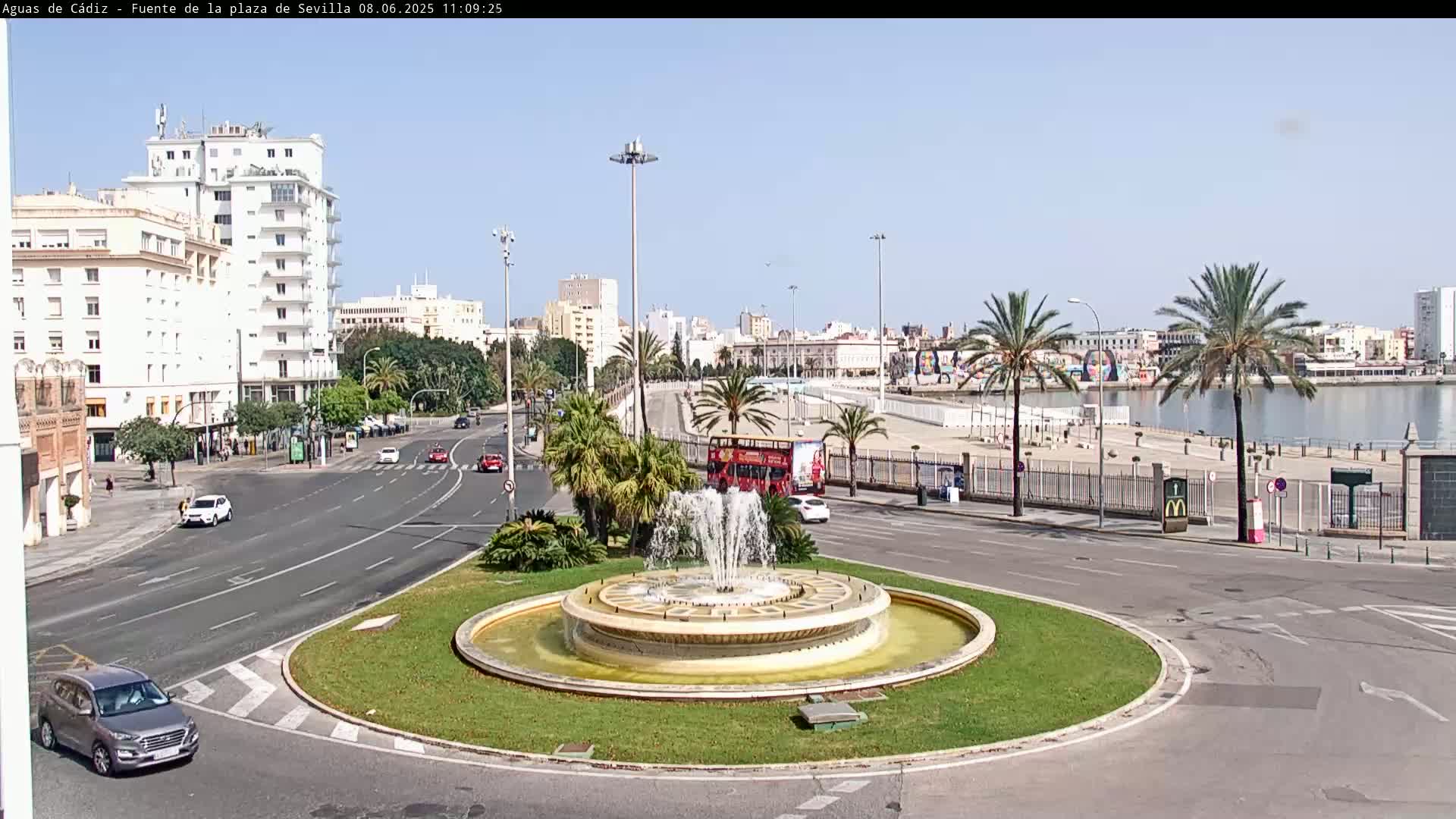 A circular fountain is at the center of a traffic circle, with city buildings and a body of water visible in the background on a sunny day.