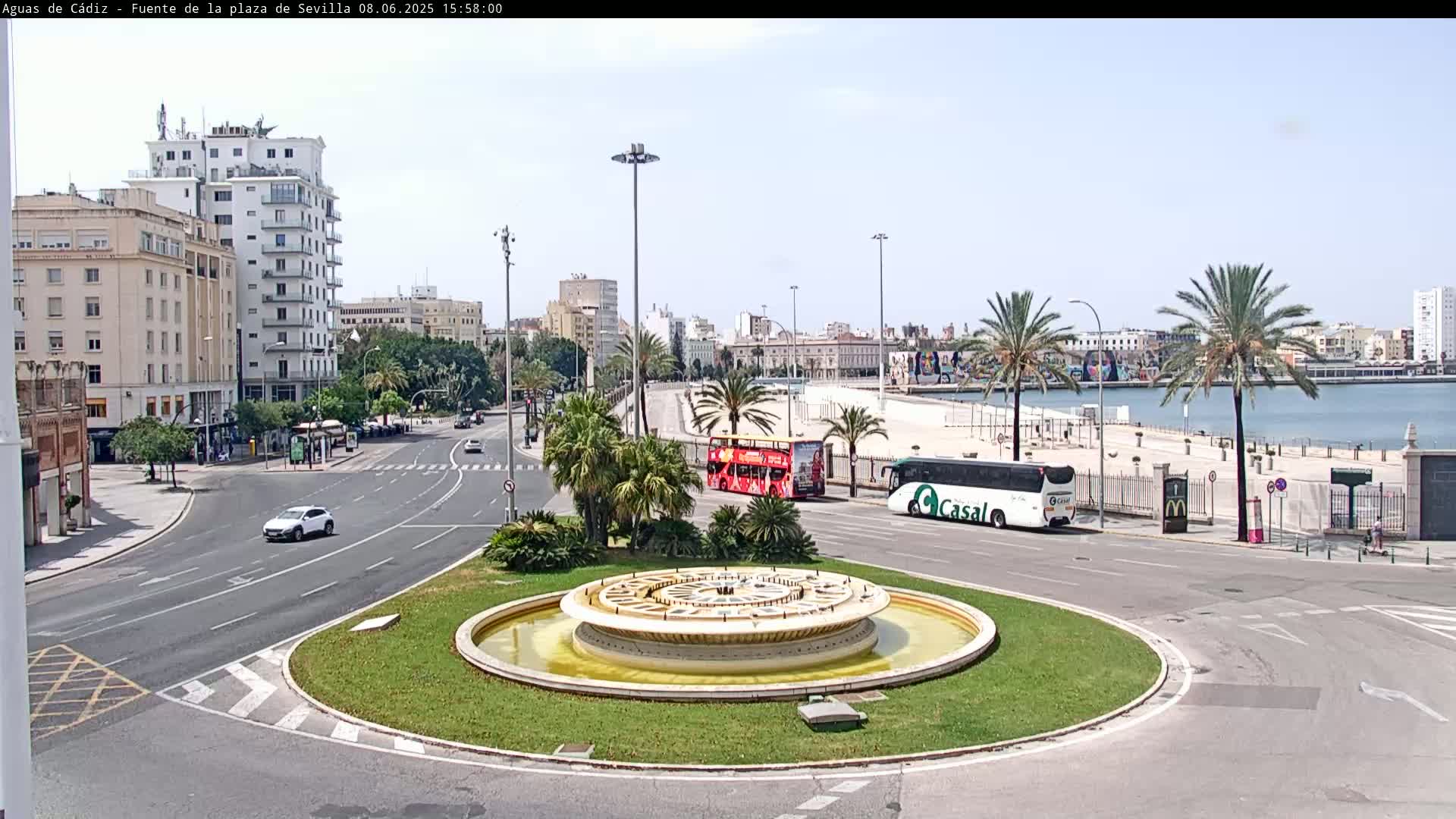A circular fountain sits in the center of a traffic circle, with buildings, palm trees, buses, and a waterfront visible in a sunny, clear day.