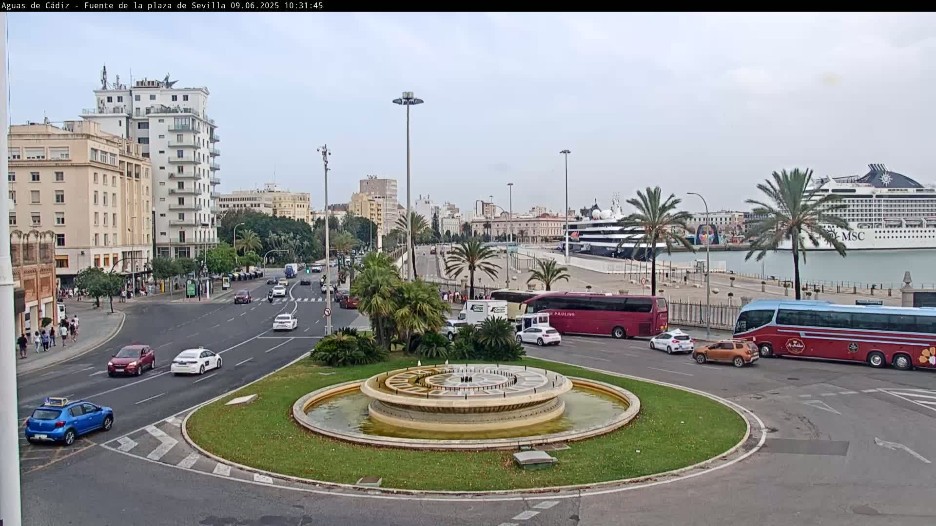 A circular fountain sits in the center of a roundabout, surrounded by city streets with cars and buses, and in the background, a harbor with docked cruise ships under a cloudy sky.