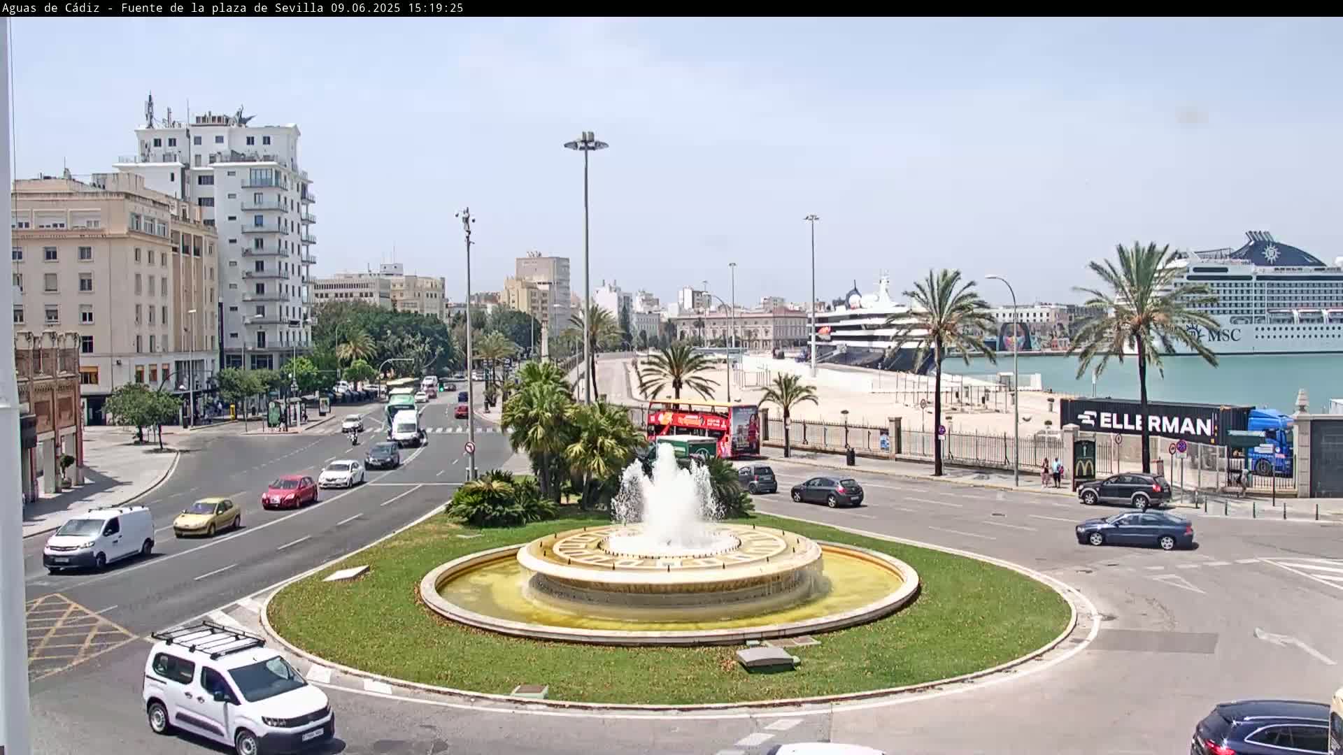 A city roundabout with a central fountain is surrounded by traffic, buildings, palm trees, and a harbor with cruise ships, all under a sunny sky.