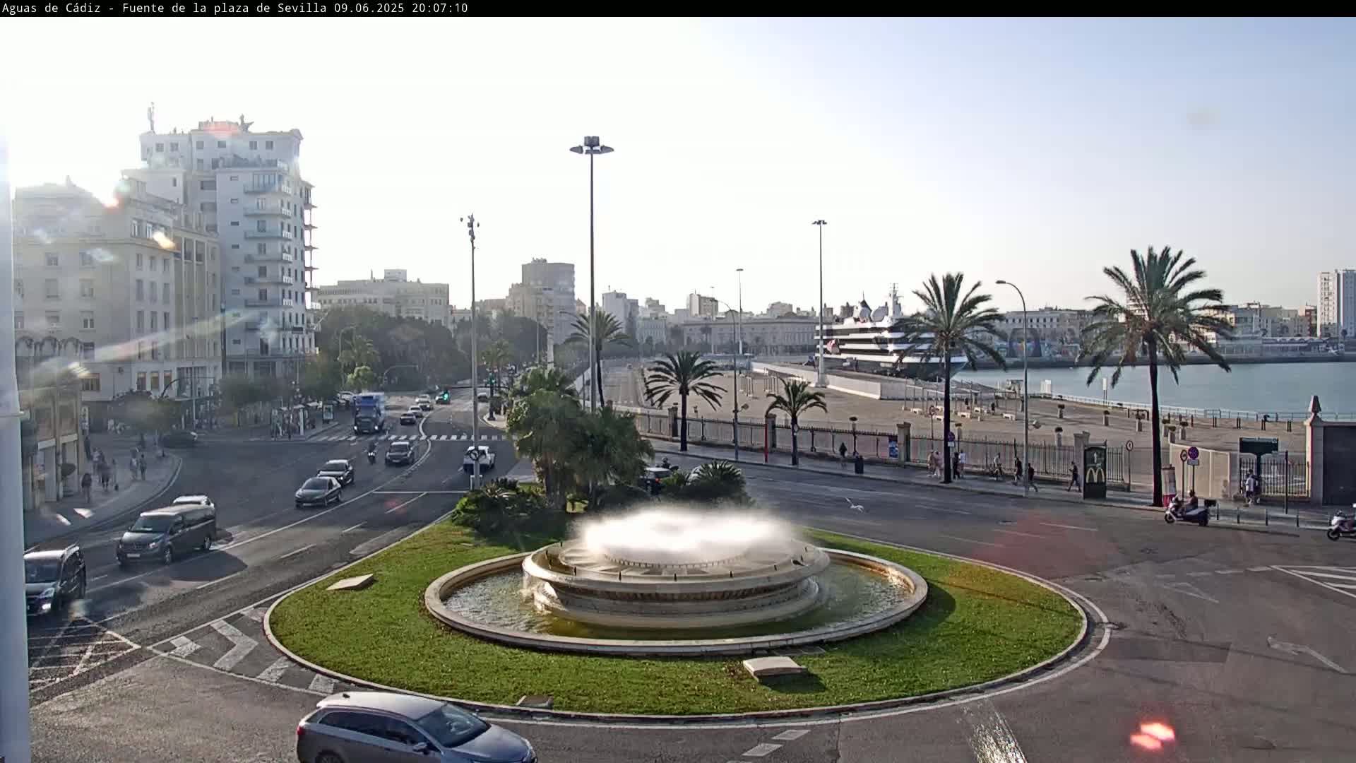 A city roundabout with a fountain in the center, surrounded by palm trees, buildings, and cars, on a partly sunny day.