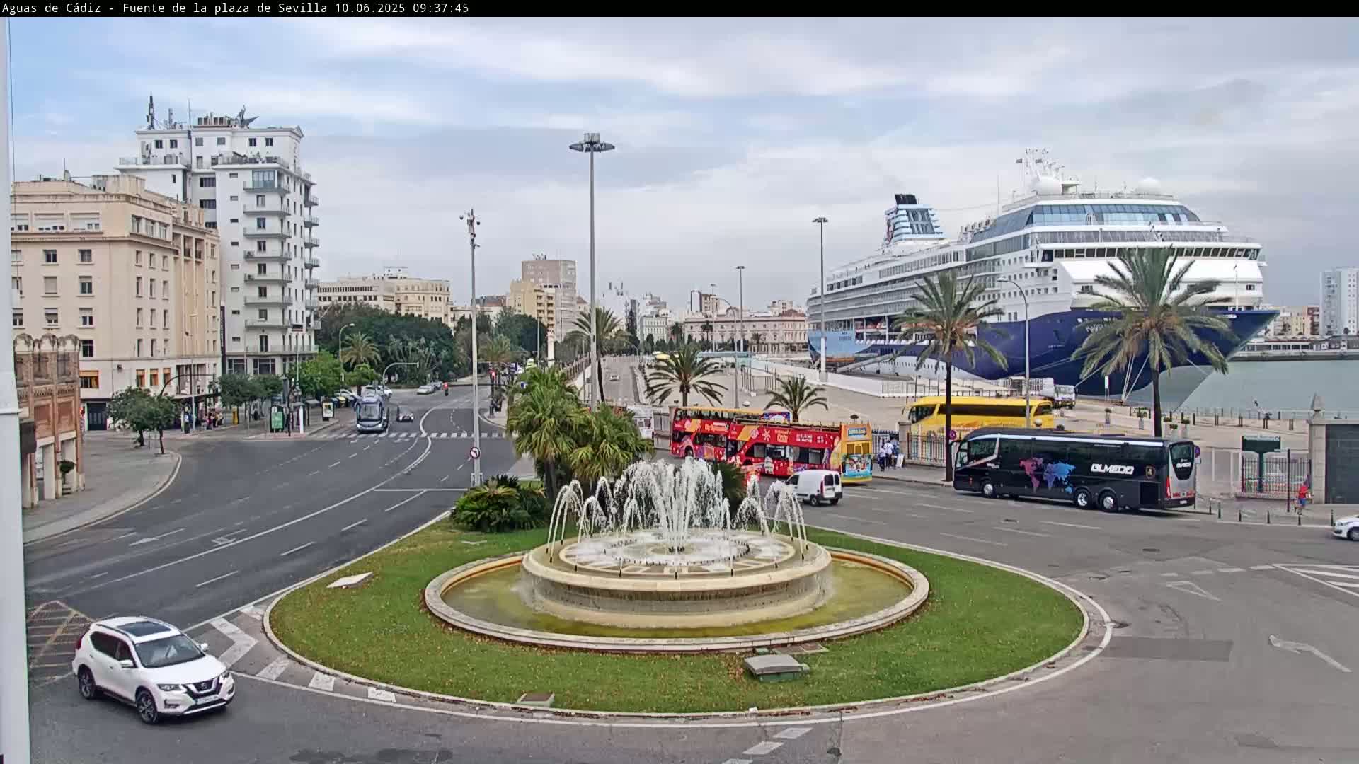 A large cruise ship is docked at a port near a city roundabout with a fountain, and several buses and cars are visible on the streets under an overcast sky.