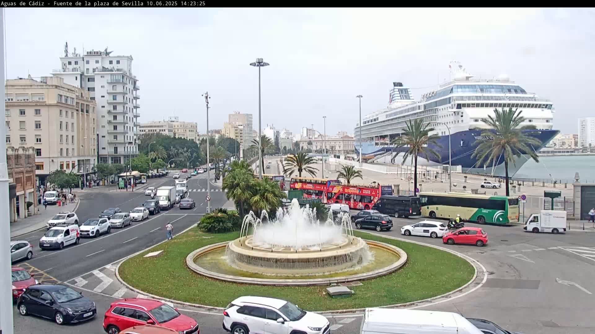 A large cruise ship is docked at a port, with a roundabout containing a fountain in the foreground, surrounded by city buildings and traffic under an overcast sky.