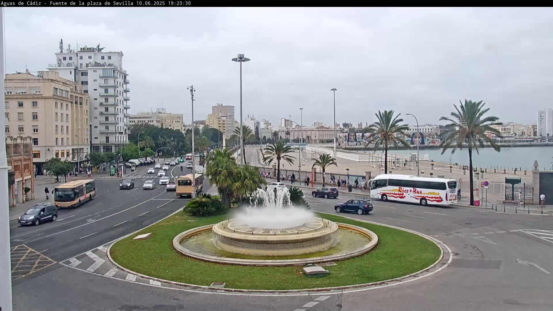 A city street scene on an overcast day features a large fountain in a roundabout, with cars and buses traveling on the road, palm trees lining the waterfront, and buildings in the background.