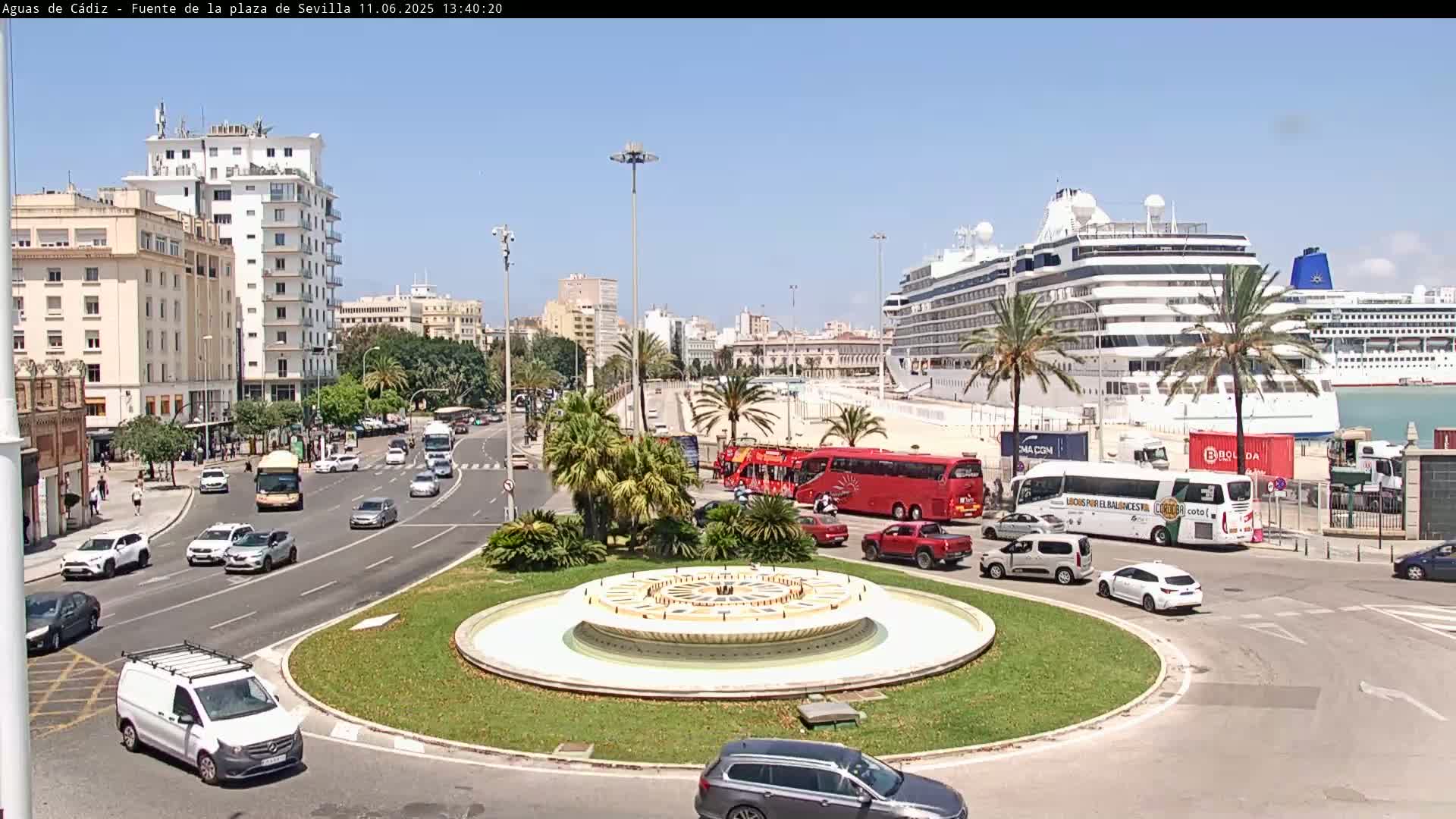 A city roundabout with a fountain in the center, surrounded by traffic including cars and buses, with a large cruise ship docked in the harbor in the background on a sunny day.