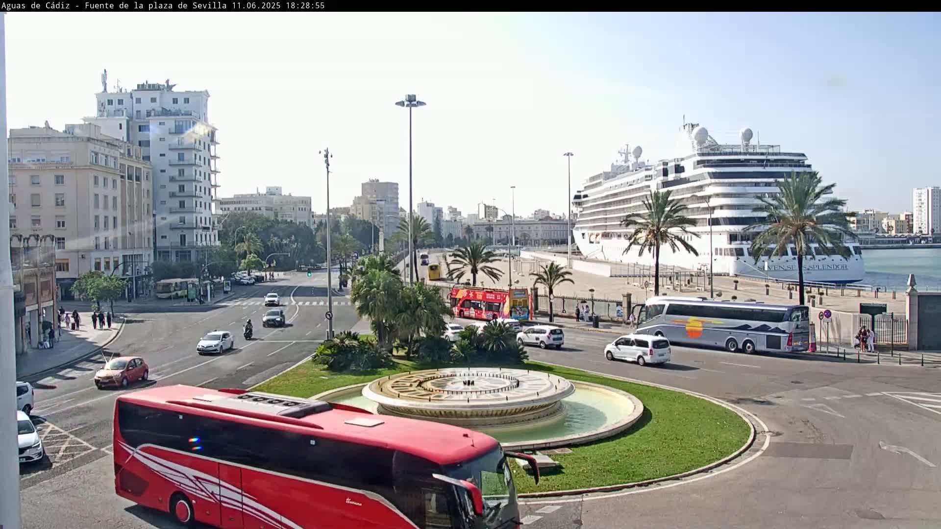 A city square with a fountain at its center, surrounded by roads with buses and cars, and a large cruise ship docked in the background on a sunny day.