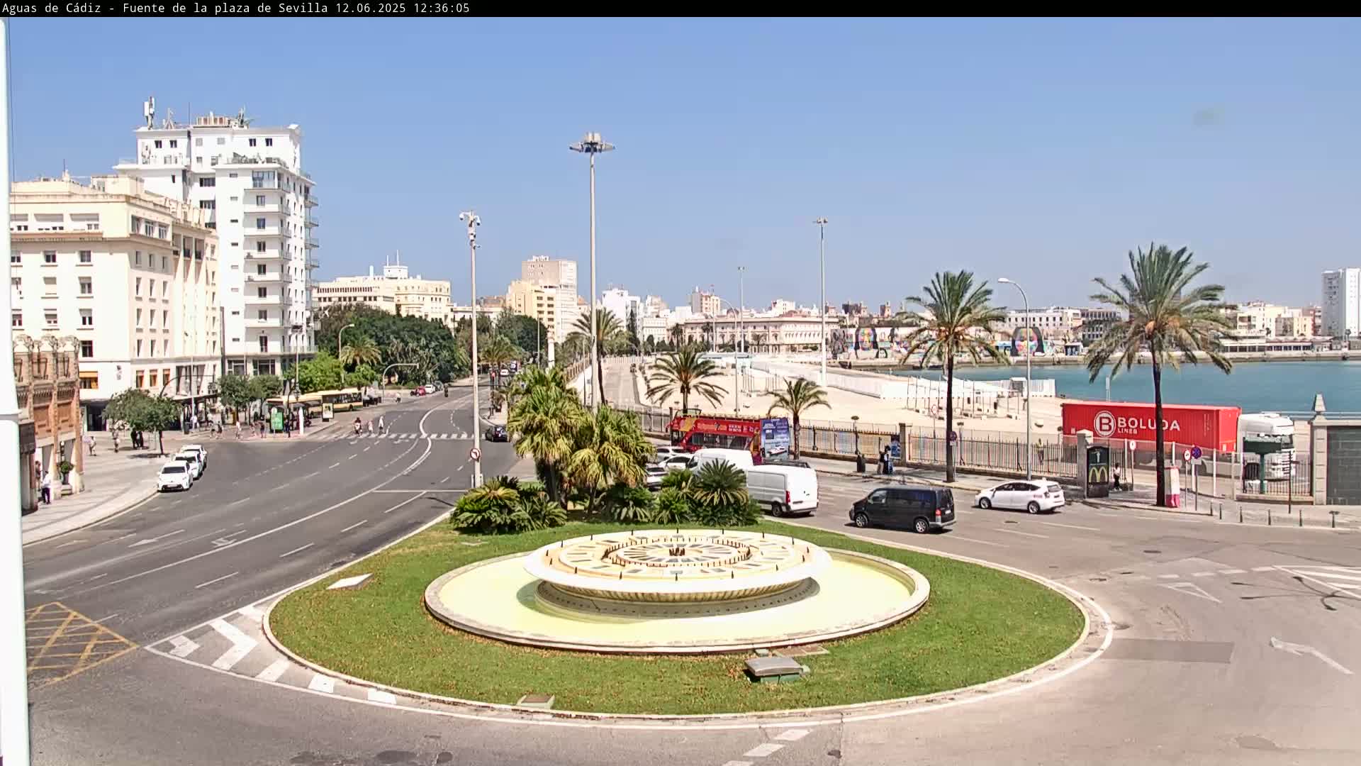 A circular fountain sits in the center of a traffic circle near a beach, with buildings, palm trees, and vehicles visible under a clear, sunny sky.