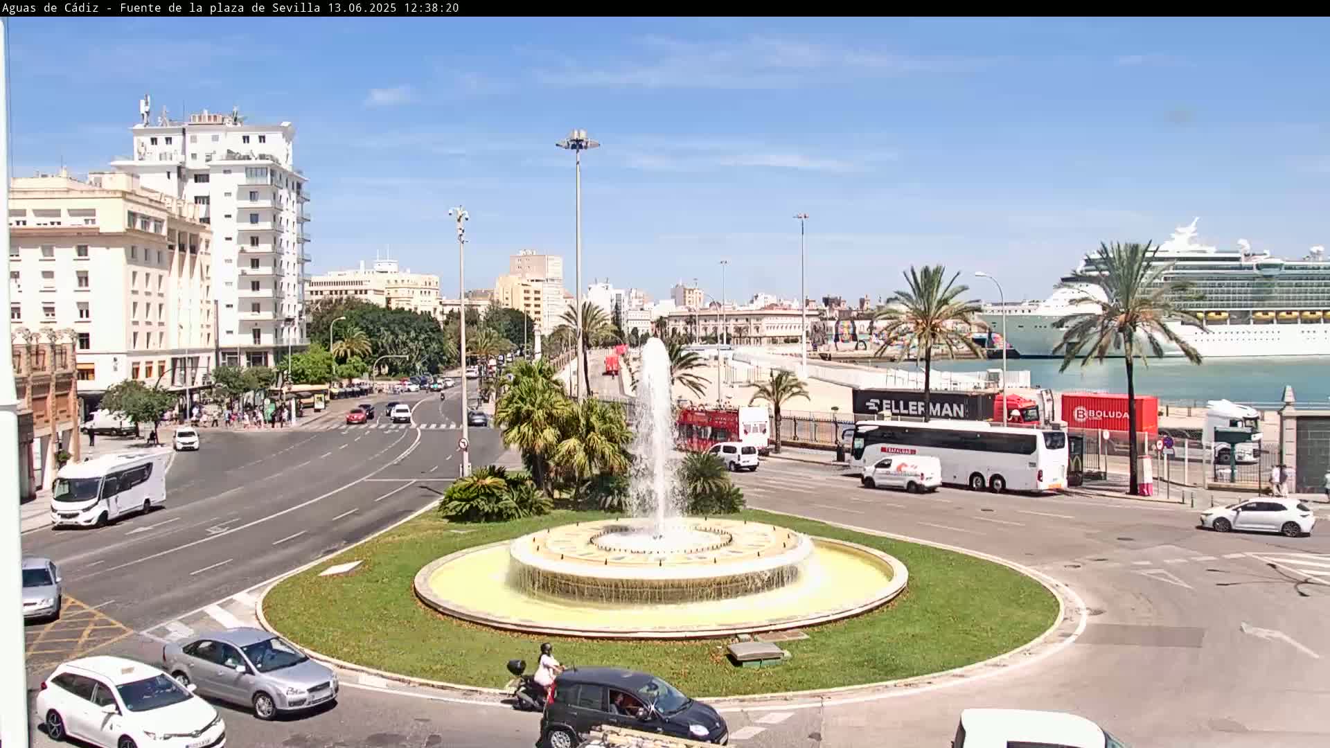 A large fountain sits in a circular traffic island surrounded by city streets, buildings, and palm trees, with a cruise ship visible in the harbor under a sunny sky.