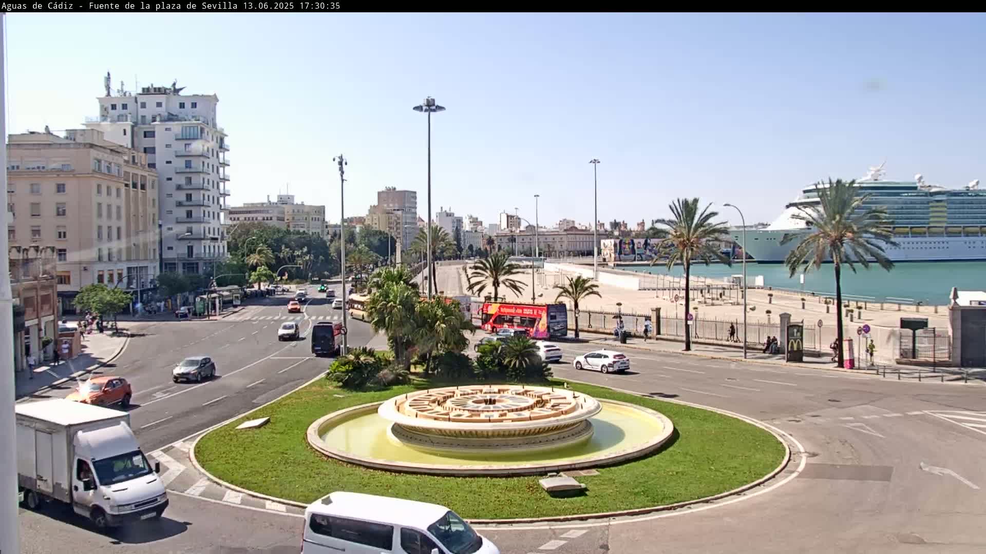 A circular fountain is in the center of a roundabout surrounded by city streets and buildings, with a large cruise ship visible in the harbor on a sunny day.