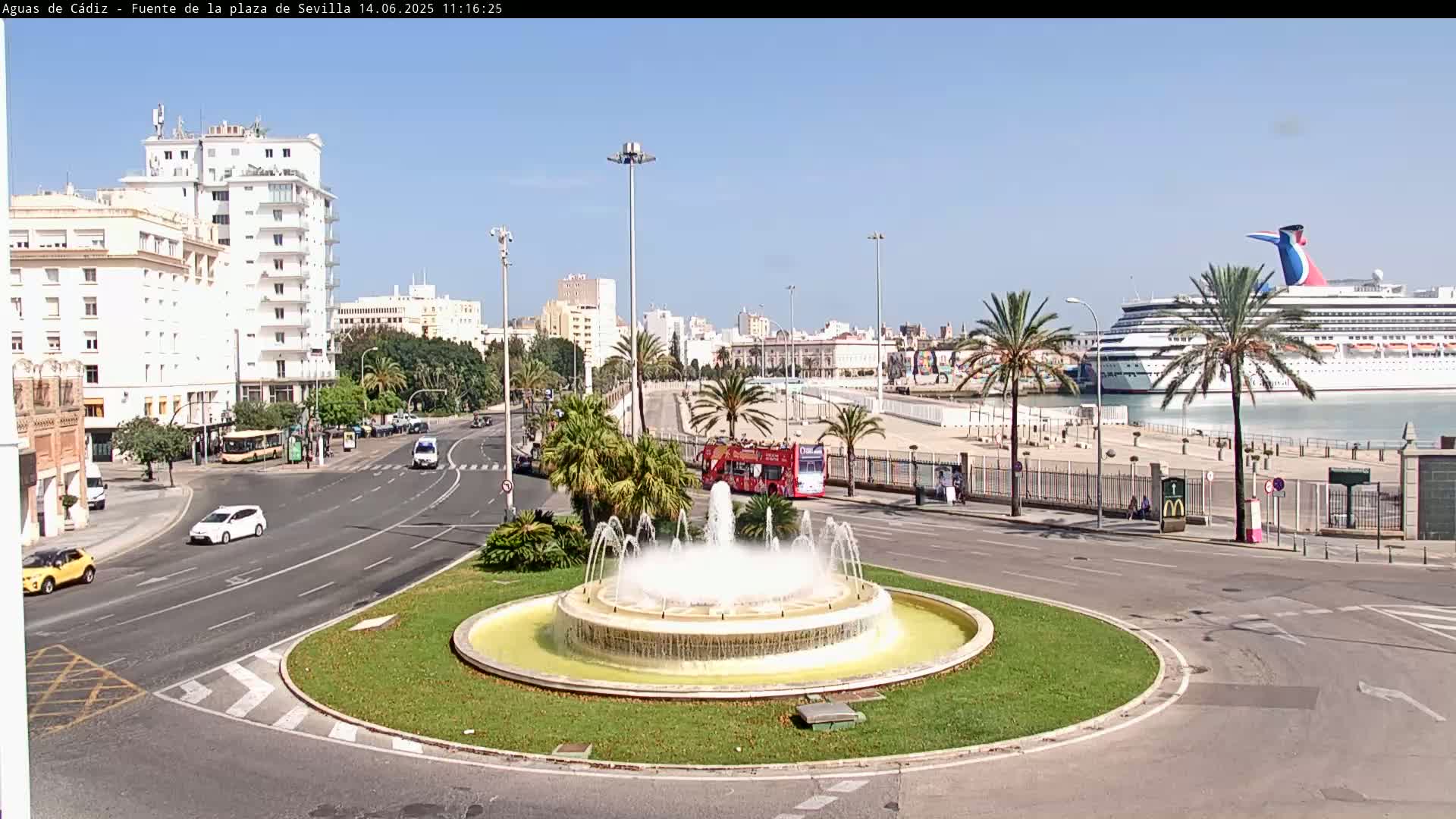 A circular fountain in a city plaza, with a large cruise ship visible in the harbor in the background on a sunny day.