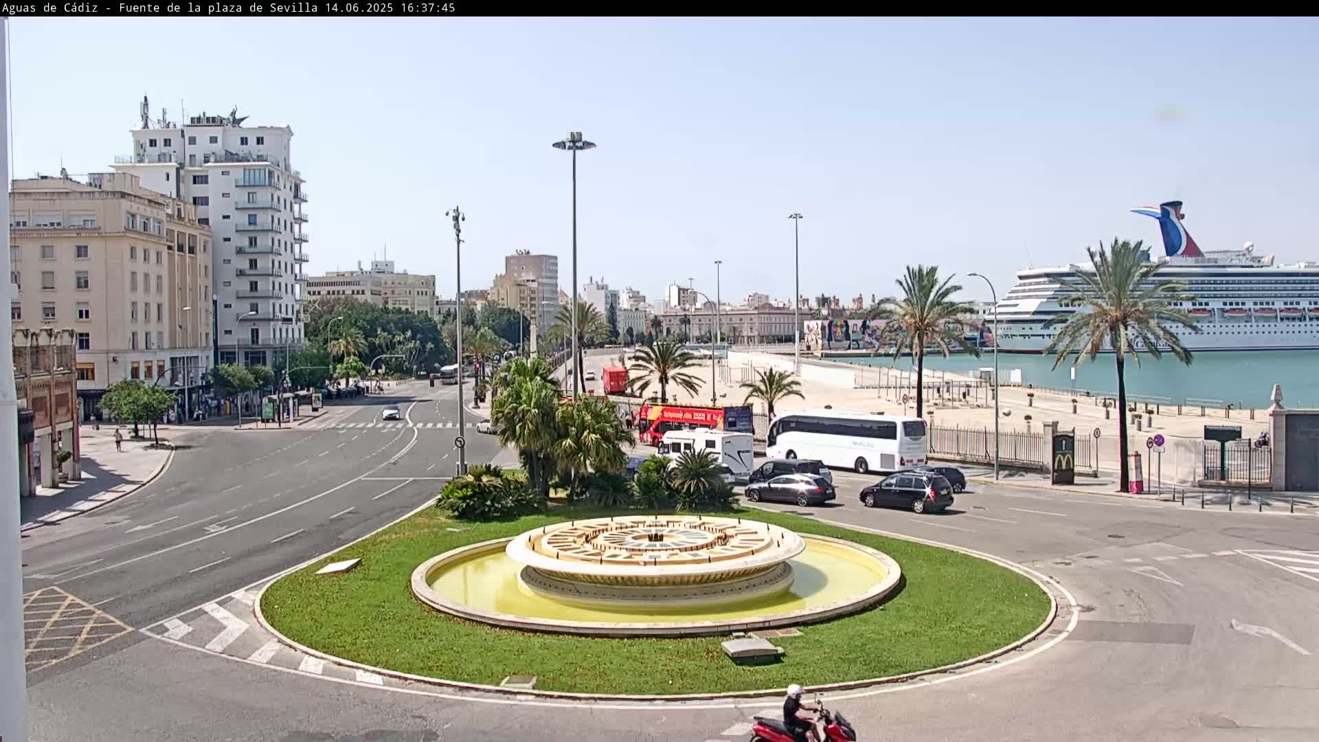 A circular fountain sits in the center of a traffic circle near a harbor where a large cruise ship is docked, under a clear sunny sky.