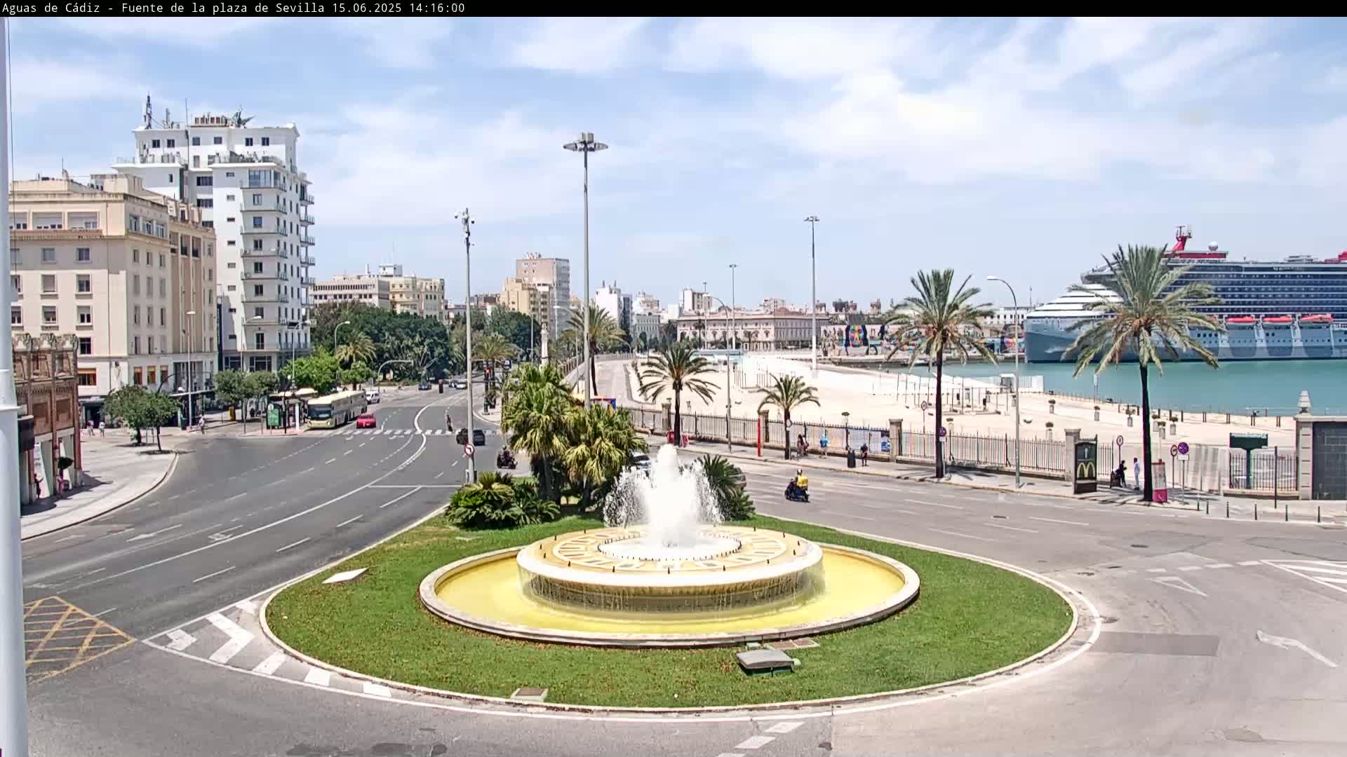 A large fountain sits in a traffic circle overlooking a harbor with a cruise ship docked on a sunny day.