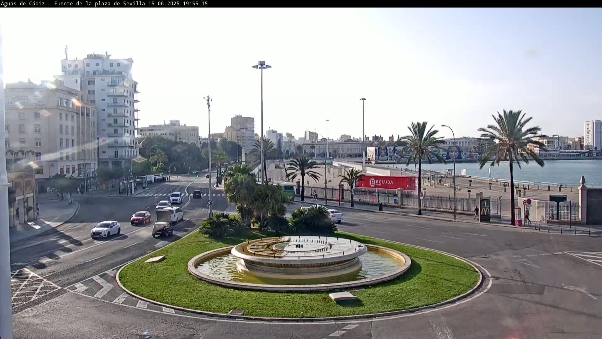 A circular fountain sits in a traffic circle overlooking a waterfront city on a sunny day.
