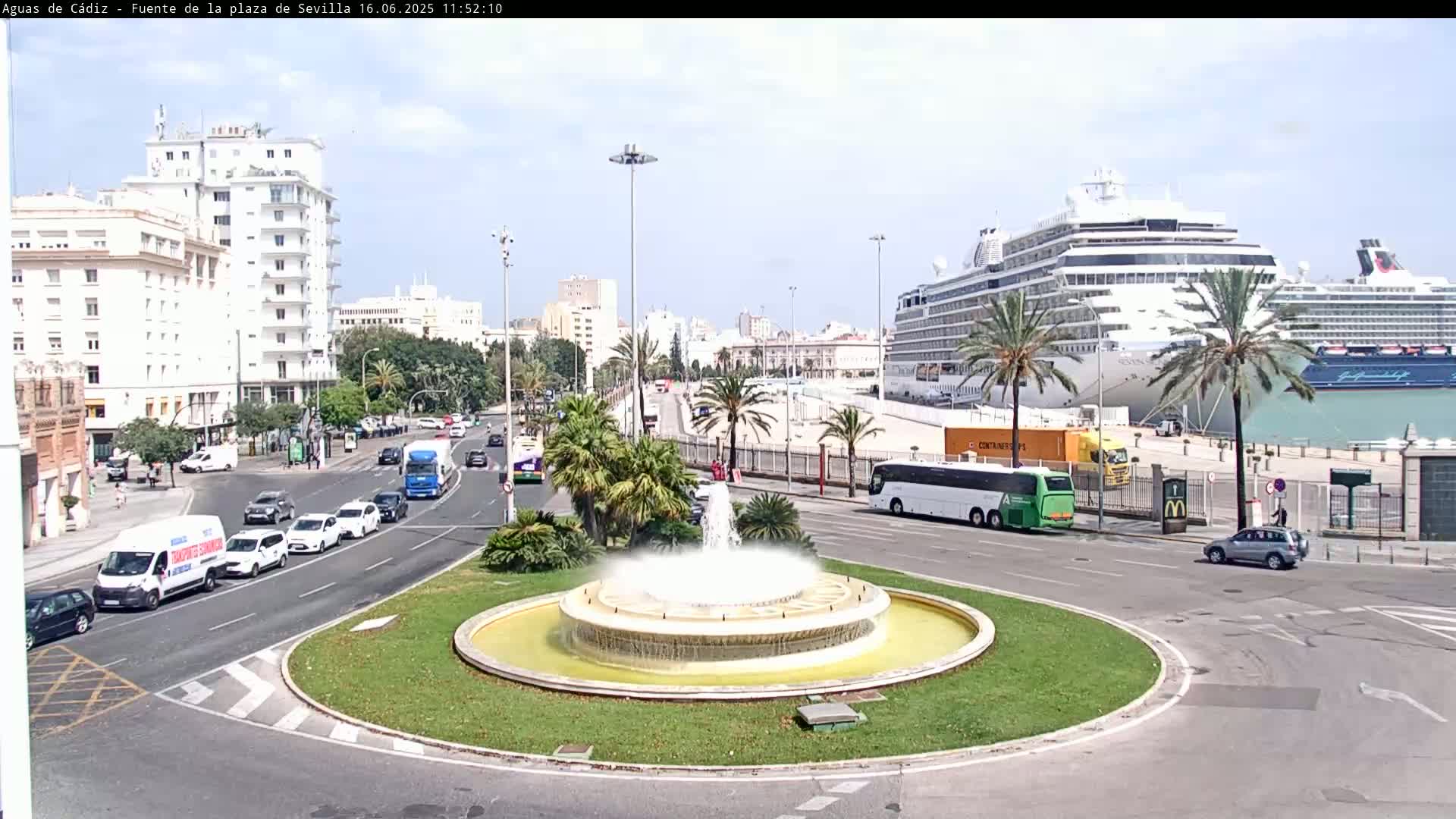 A circular fountain sits in the center of a traffic circle, with city streets, buildings, and two large cruise ships visible in the background on a sunny day.