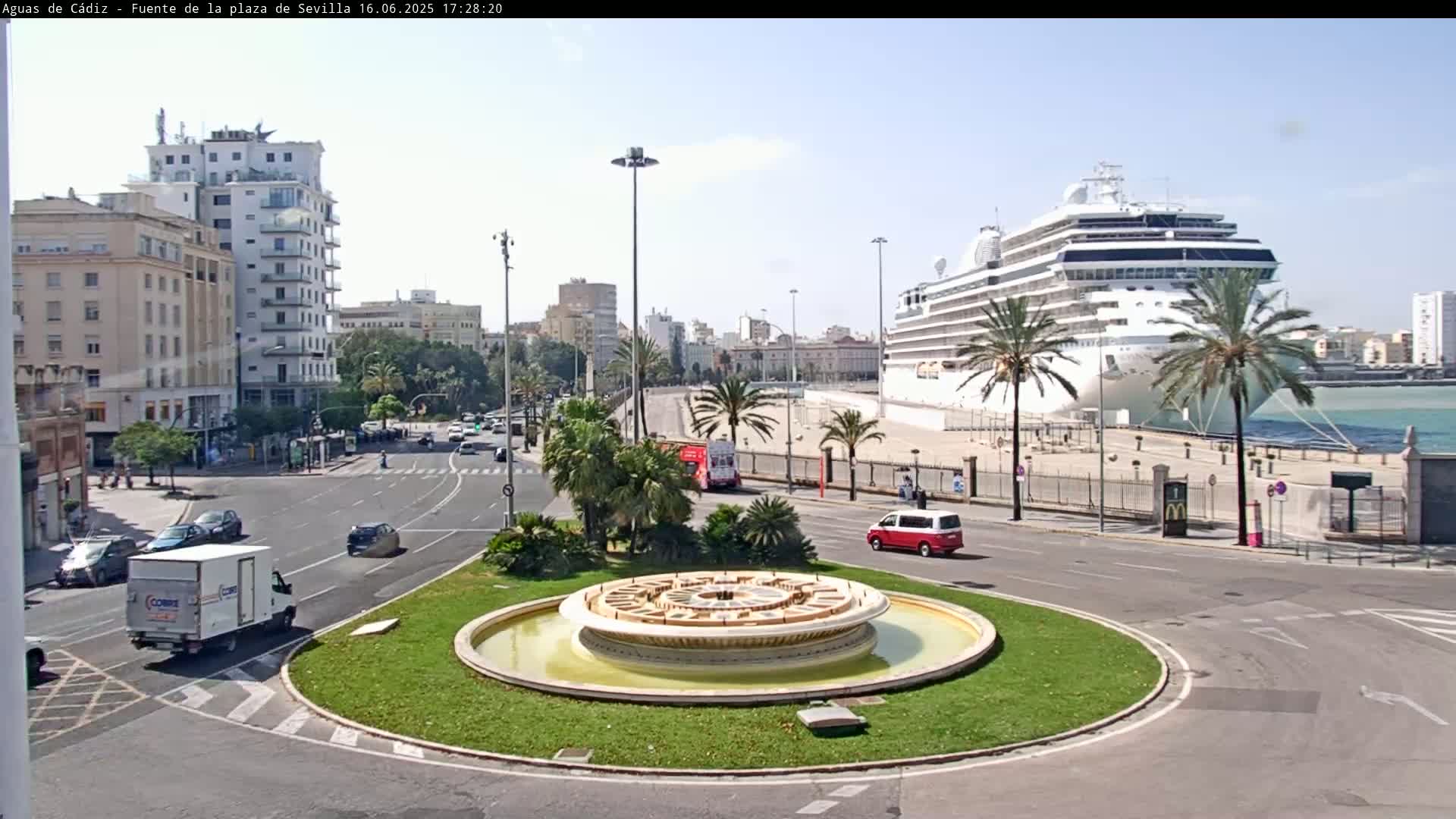 A large cruise ship is docked at a port near a city with a circular fountain in the foreground, under a sunny sky.