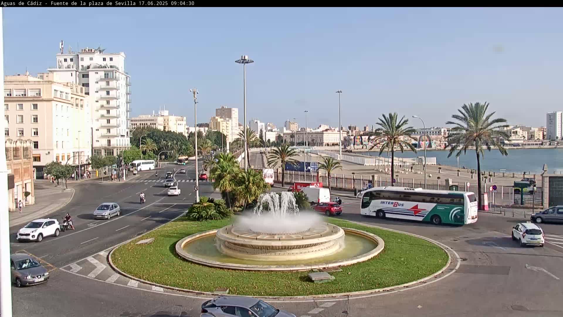 A city roundabout with a large fountain in the center, surrounded by palm trees, buildings, and cars, under a clear sky.