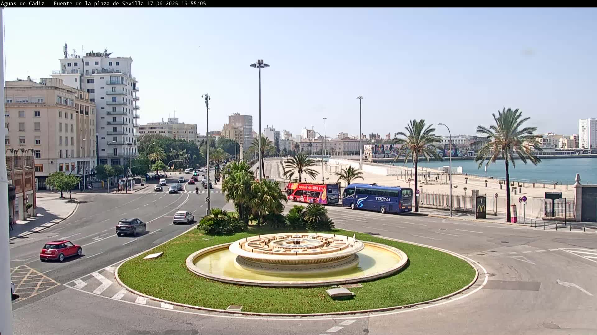 A traffic circle with a fountain in the center is surrounded by city streets and buildings, with palm trees lining a waterfront in the background under a sunny sky.