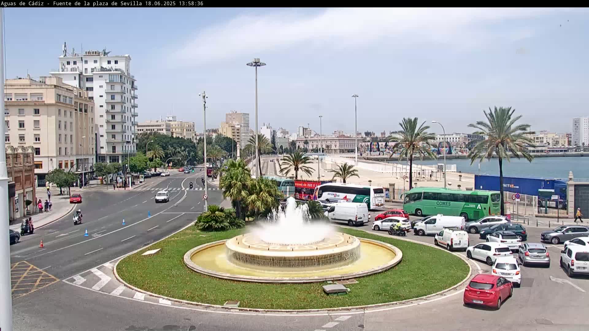 A city roundabout with a fountain in the center, surrounded by traffic including buses and cars, under a mostly clear sky.