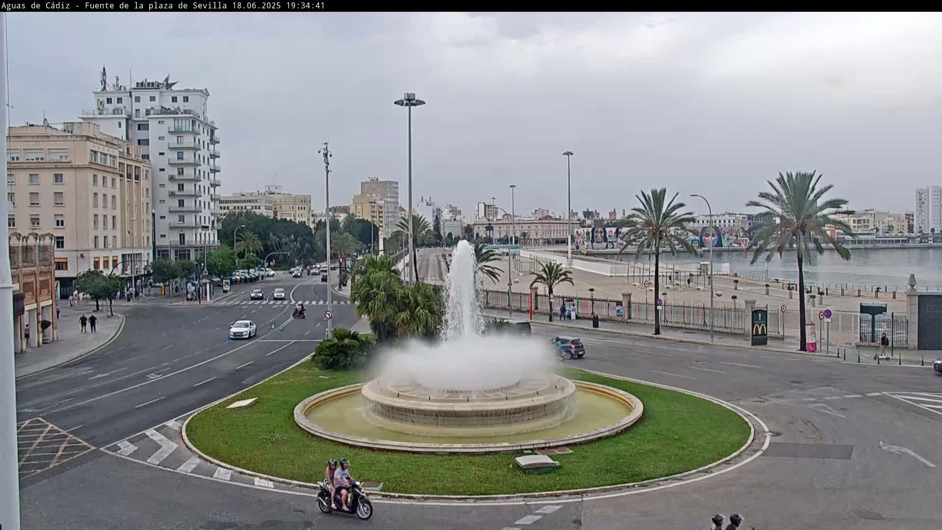 A large fountain in a city roundabout sprays water on an overcast day, with buildings, palm trees, and a waterfront visible in the background.