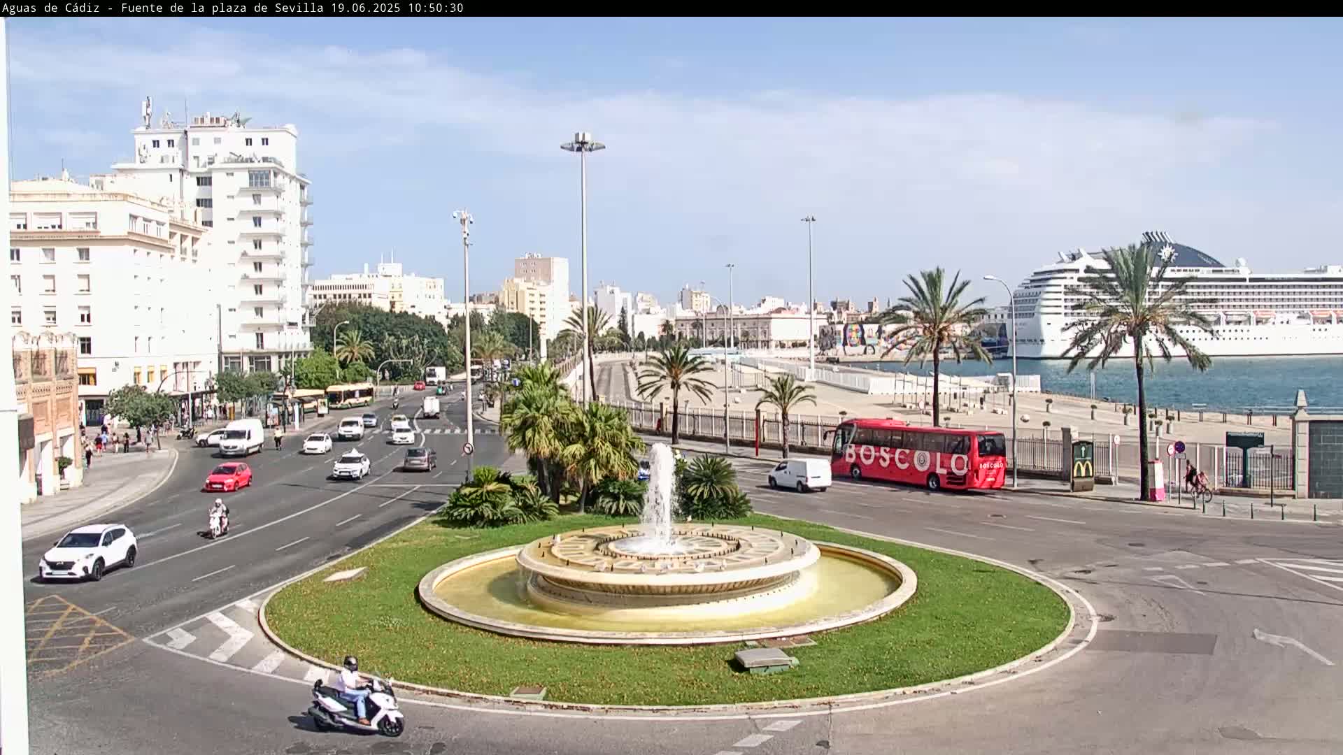 A city roundabout with a central fountain features cars and a red bus on a sunny day, with a large cruise ship visible in the harbor in the background.