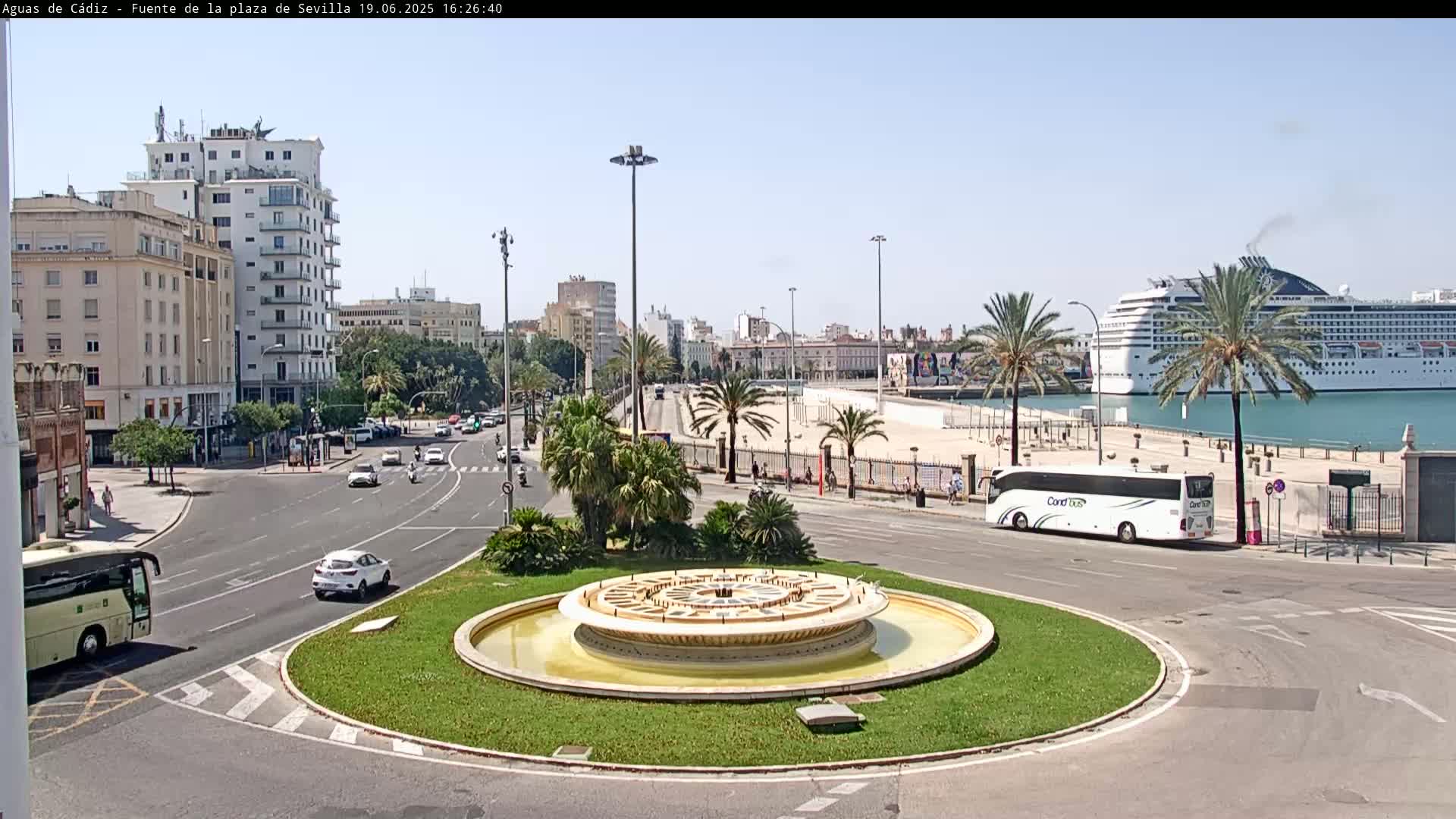 A roundabout with a central fountain is situated near a harbor where a cruise ship is docked, under a clear sunny sky.